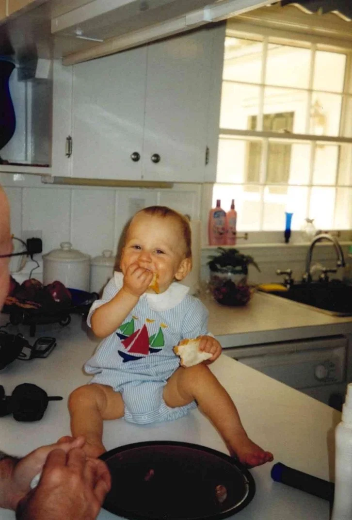 A young child with blond hair sitting on a kitchen counter, eating a snack and smiling at the camera, with kitchen cabinets, a window, and various kitchen items in the background.