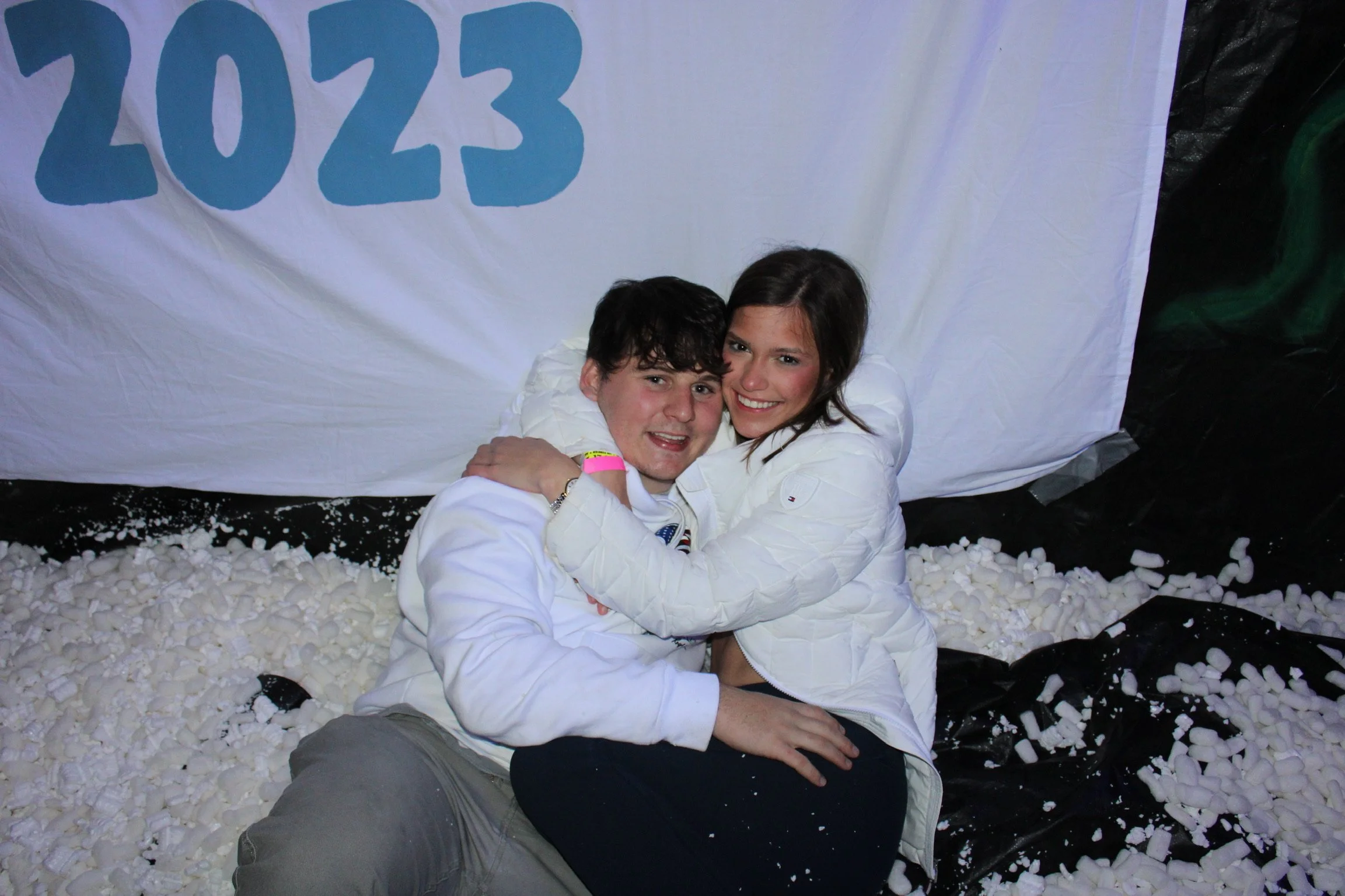 A young man and woman sitting together, smiling and hugging on foam peanuts, with a white backdrop with the year 2023 written on it in blue.