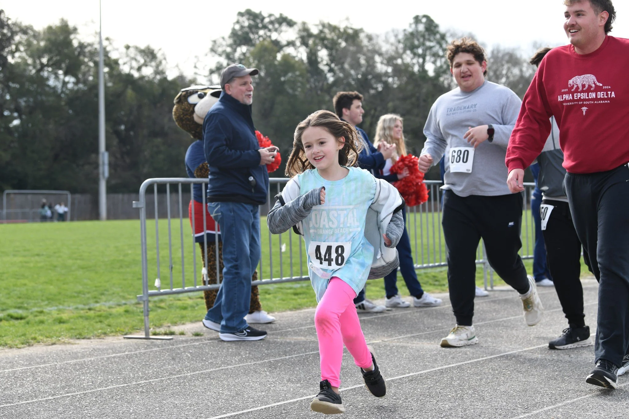 Young girl running in a race with others, wearing a race bib number 448, on a track field with spectators in background, some clapping and cheering.