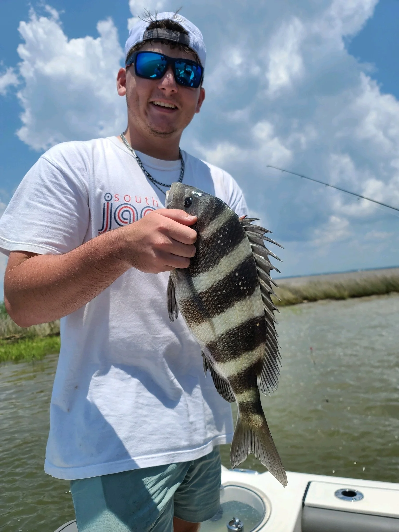 Young man wearing sunglasses, a backwards cap, a white t-shirt, and shorts, holding a large fish with vertical black and white stripes, standing on a boat in a waterway with grassy banks and a partly cloudy sky.