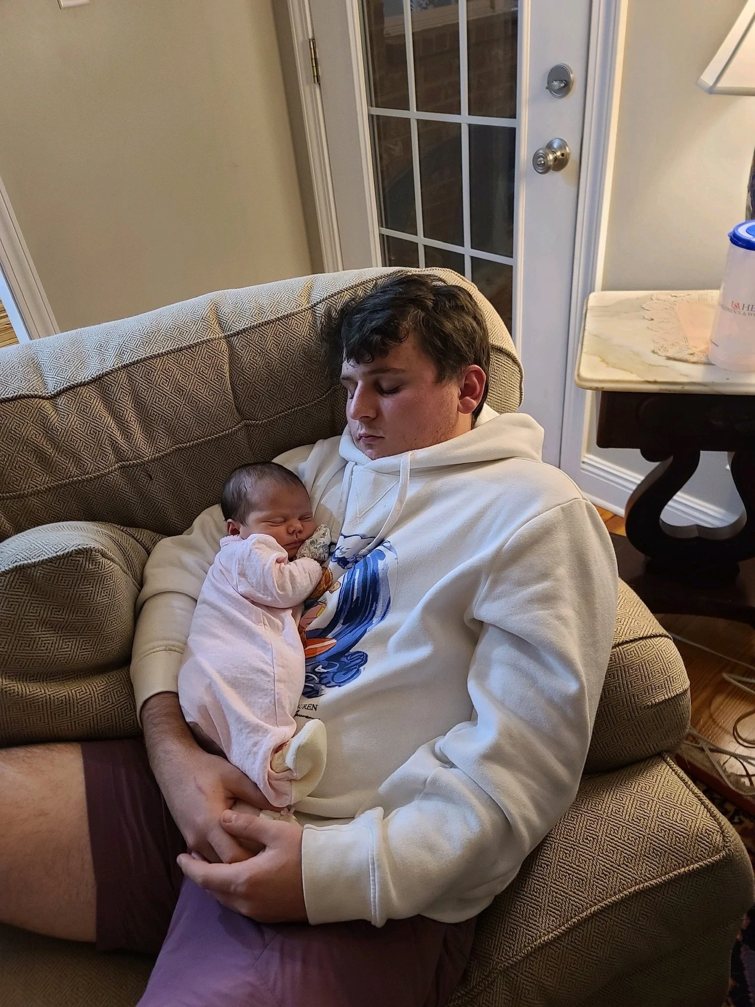 A young man sitting on a beige couch, holding a sleeping baby on his lap, with a door with glass panels in the background.