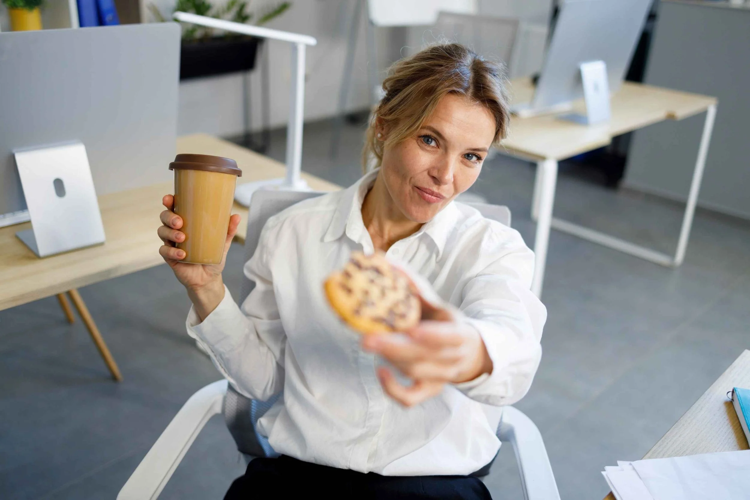 woman sitting at a desk in an office, holding a coffee cup and offering a cookie toward the camera.