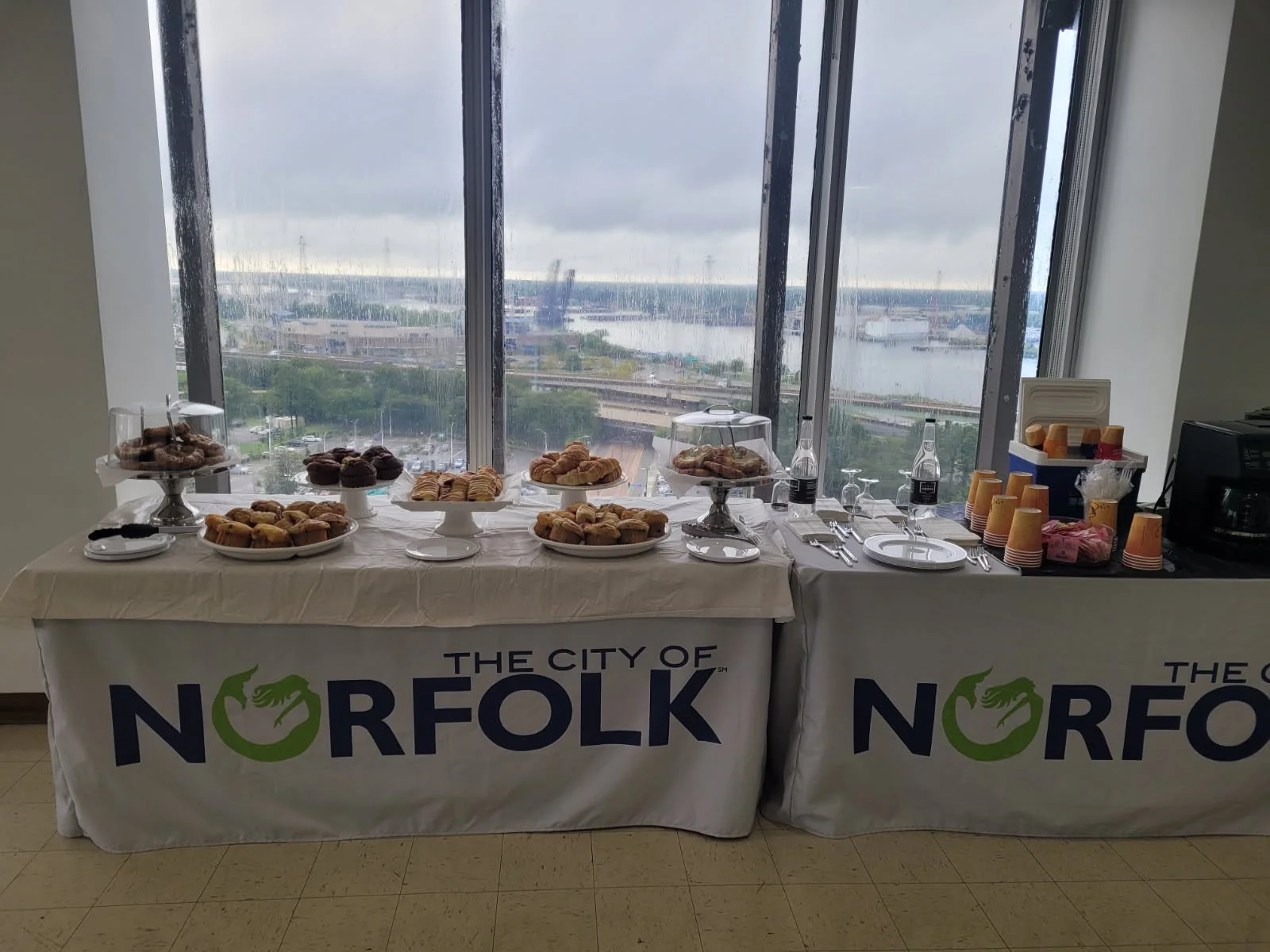 A breakfast buffet table with assorted pastries and drinks, set up in front of large windows overlooking a cityscape and a river, with cloudy skies.