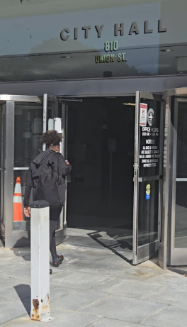 Person in black jacket and gray pants entering City Hall through glass doors with orange traffic cone nearby.
