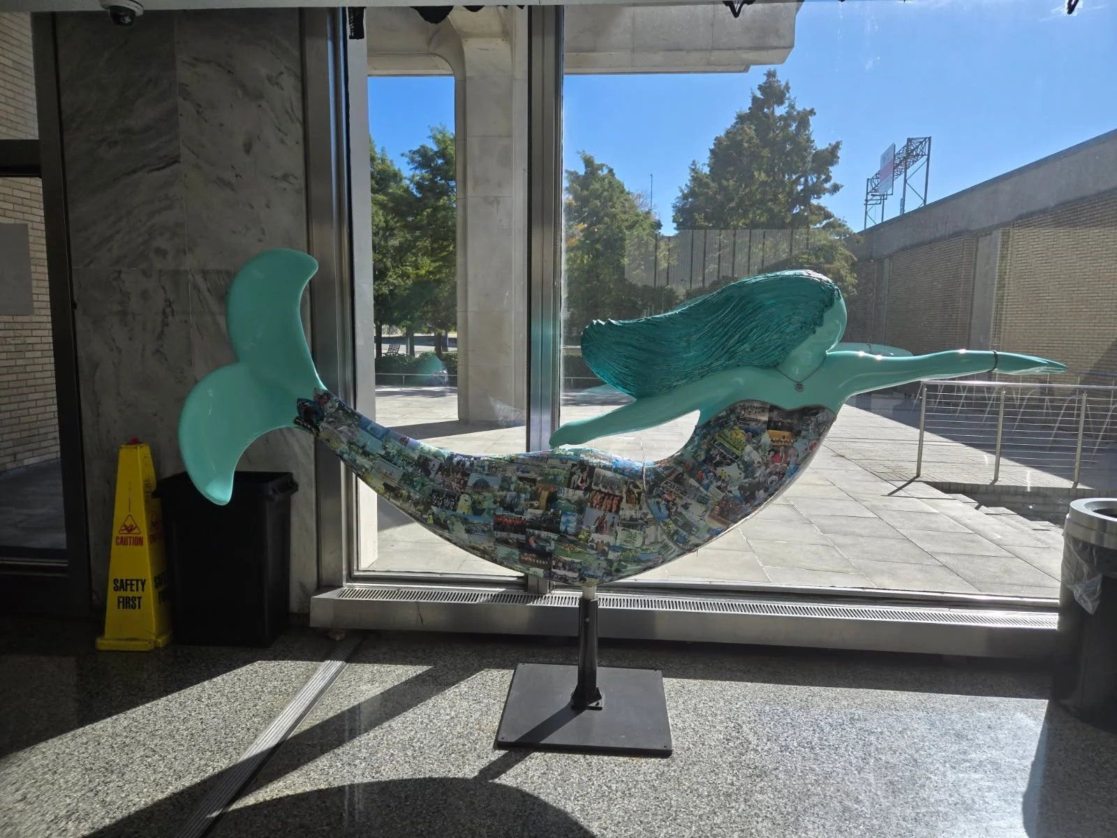 A colorful sculpture of a mermaid with a long, flowing tail and hair, positioned on a stand inside near a door with glass panels, outside visible through the glass is a sunny plaza with trees, a fence, and a brick building.