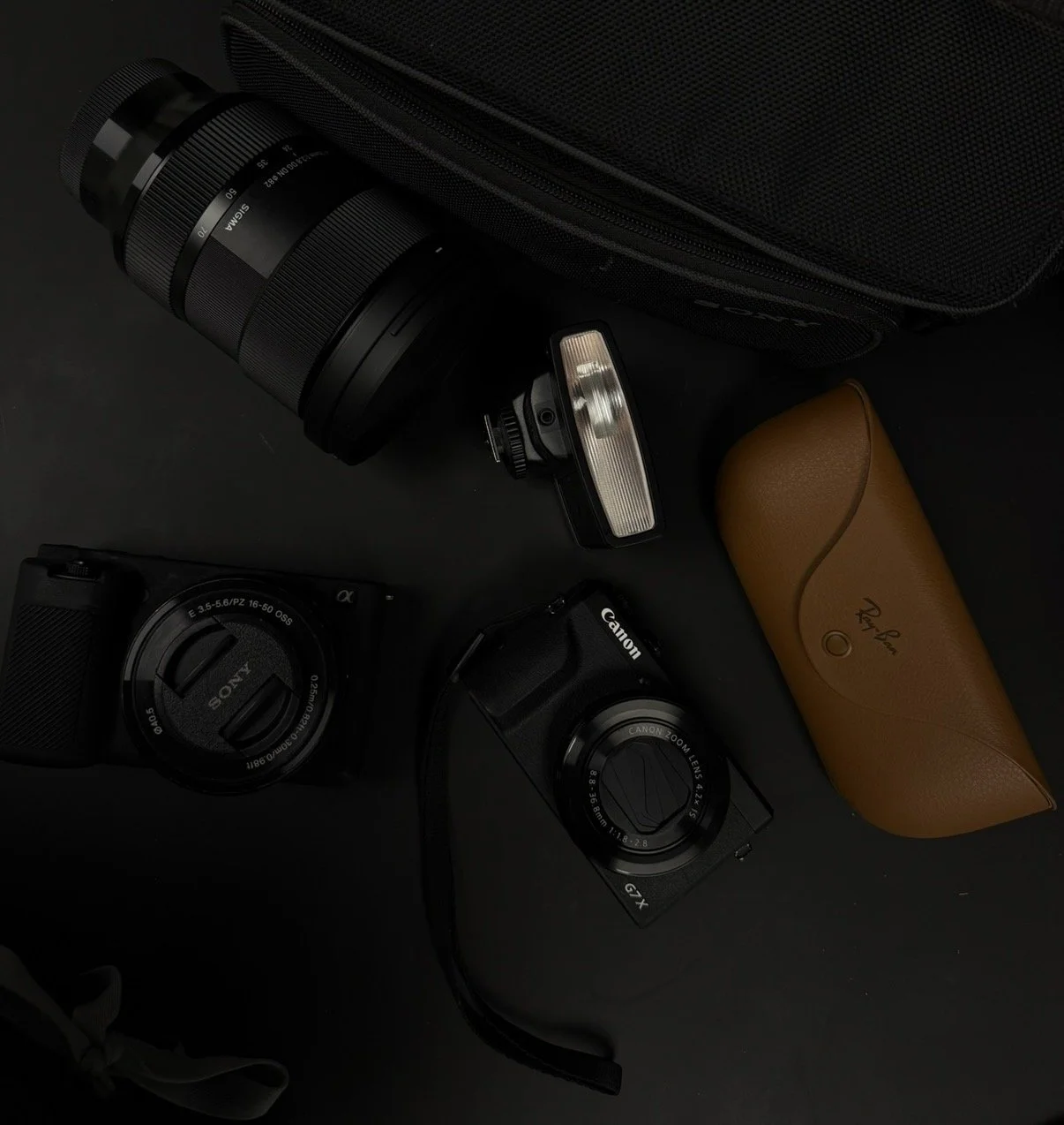 A collection of cameras, a camera flash, and accessories on a black surface, including a Sony ZV-E10 camera, a Canon G7X camera, Ray Ban Meta glasses, and a telephoto lens