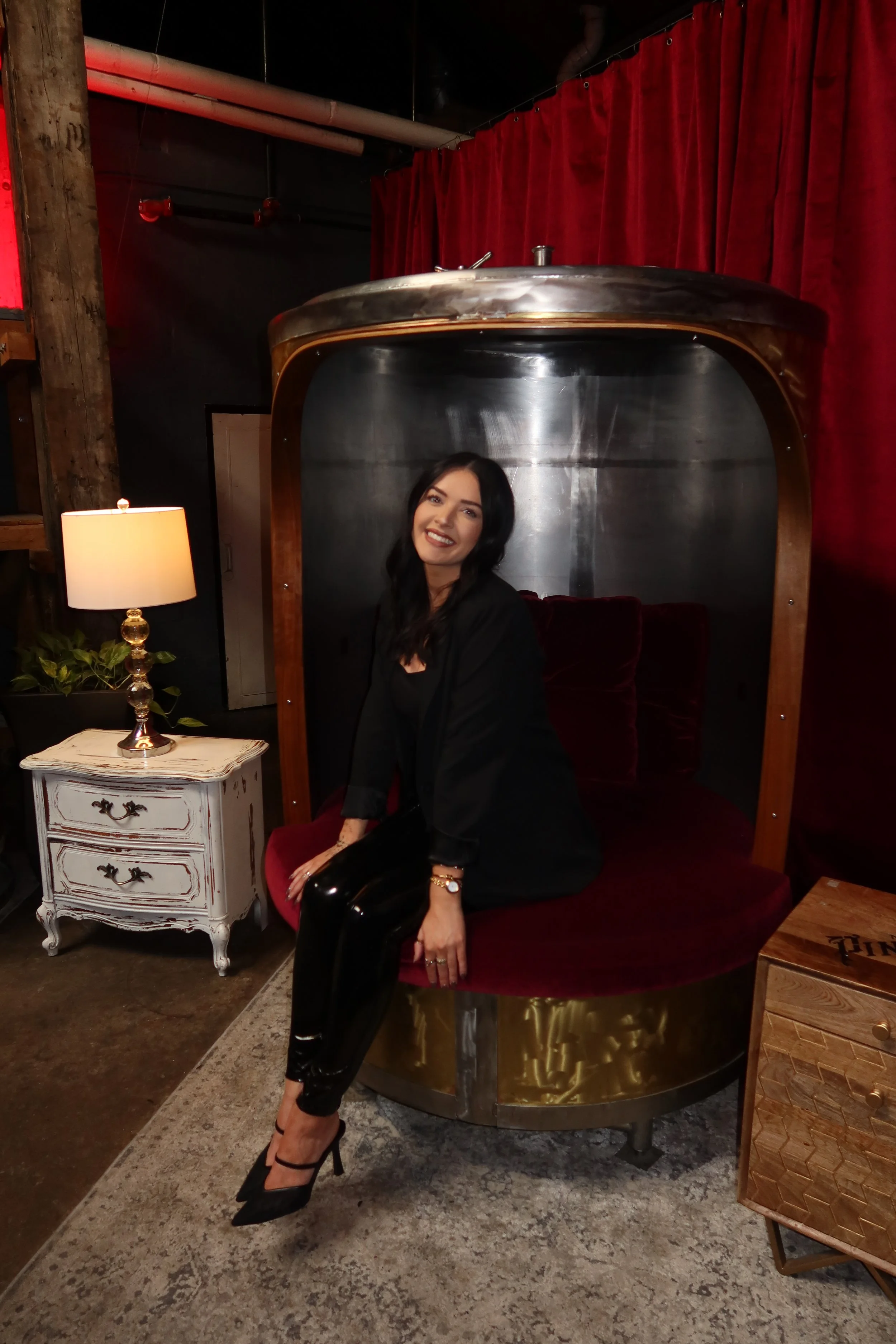 A social media business woman with black hair and a black outfit sitting inside a vintage-style, velvet-cushioned pod with a curved metallic exterior, in a room with red curtains and antique furniture.