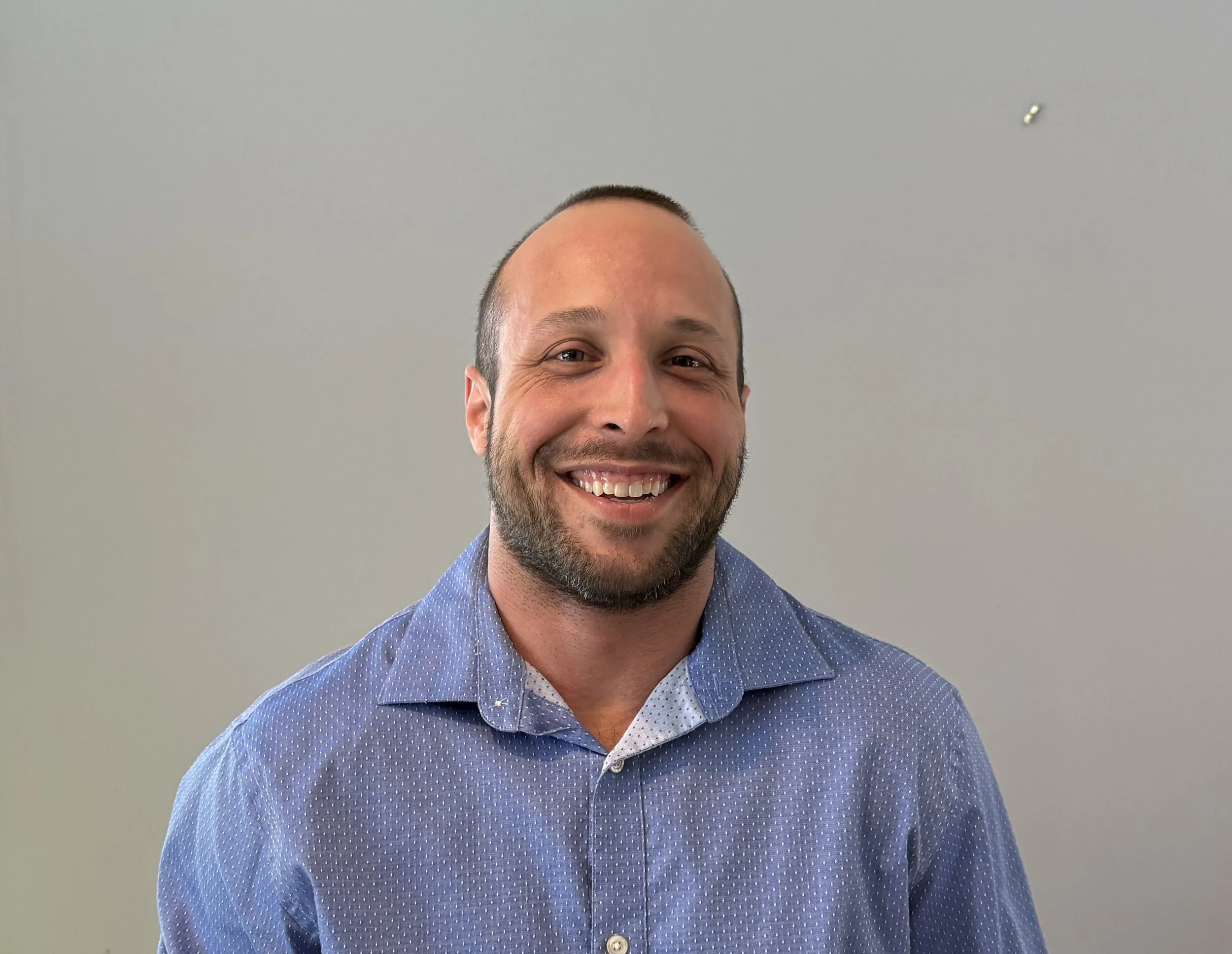A smiling man with a beard and close-cropped hair wearing a light blue, patterned dress shirt, standing against a plain gray wall.