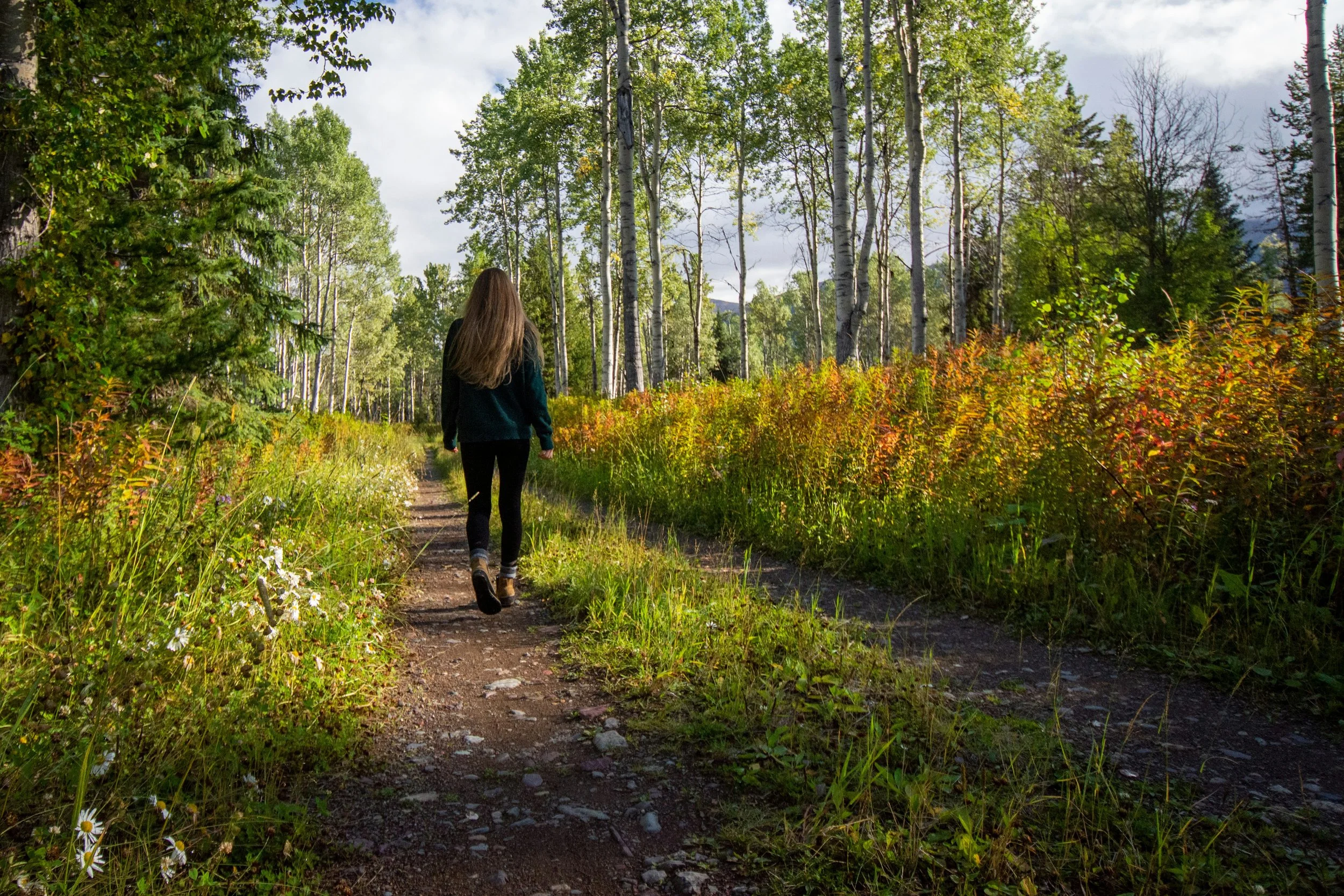 A woman with long brown hair walking on a dirt trail through a lush green forest with tall trees and bushes.