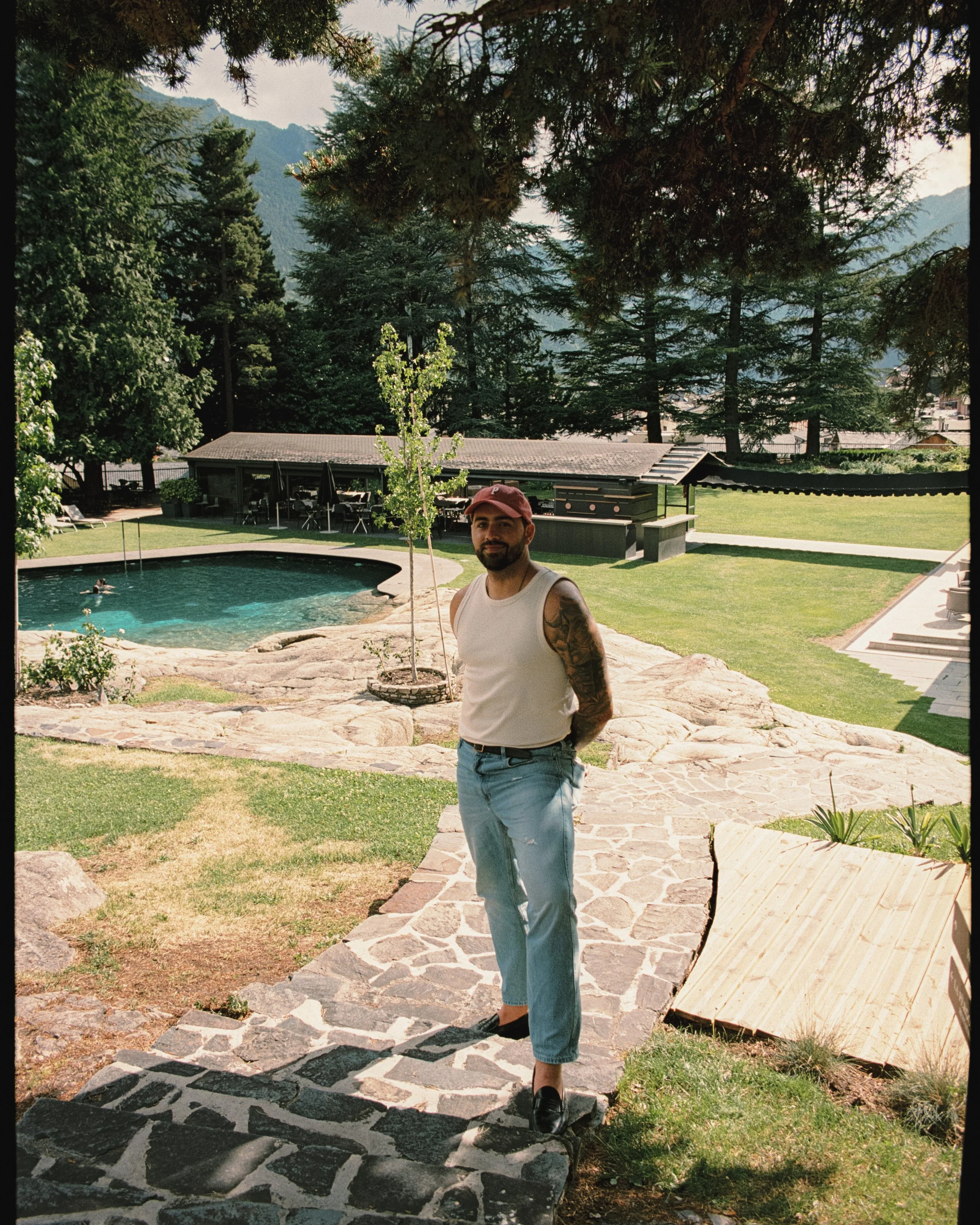 Hombre con camiseta blanca, jeans cortos y gorra roja en un jardín con piscina y árboles en el fondo