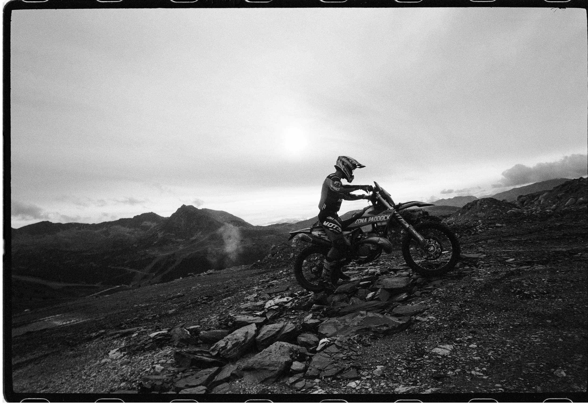 Persona montando una motocicleta en terrenos rocosos con montañas al fondo en una fotografía en blanco y negro.