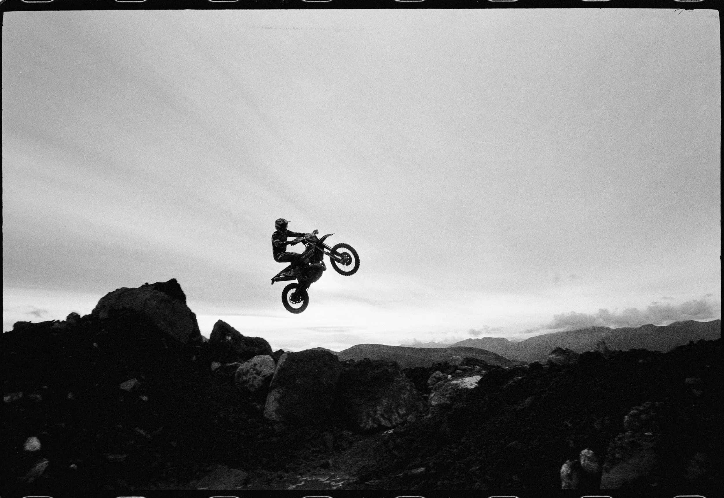 Una persona en una motocicleta haciendo truco en el aire en un paisaje con rocas y montañas al fondo, en blanco y negro.