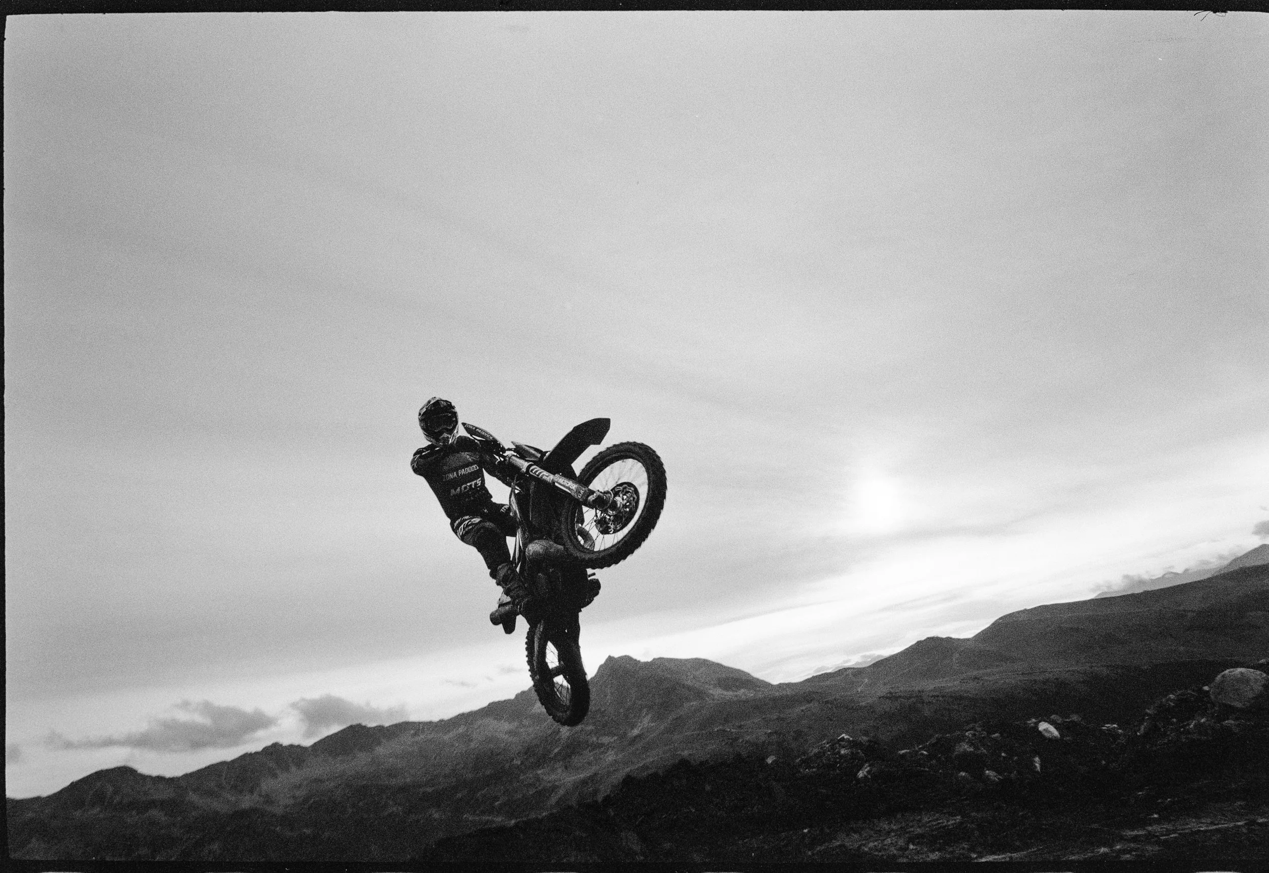 Un ciclista realizando un salto en una montaña, en un paisaje natural con montañas al fondo, en blanco y negro.