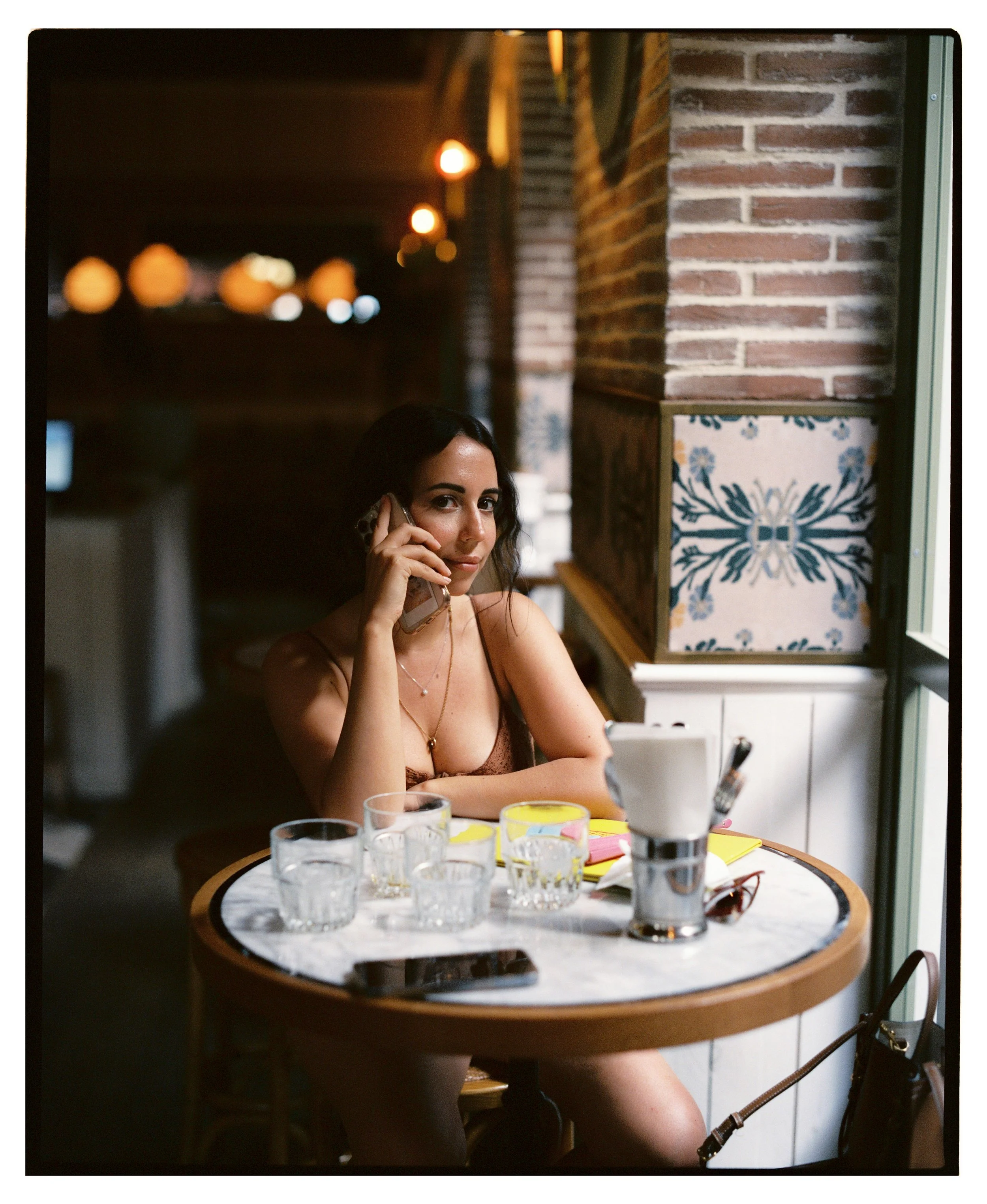 Mujer sentada en una mesa de restaurante, hablando por teléfono, con vasos de agua y algunos objetos en la mesa, frente a una ventana.