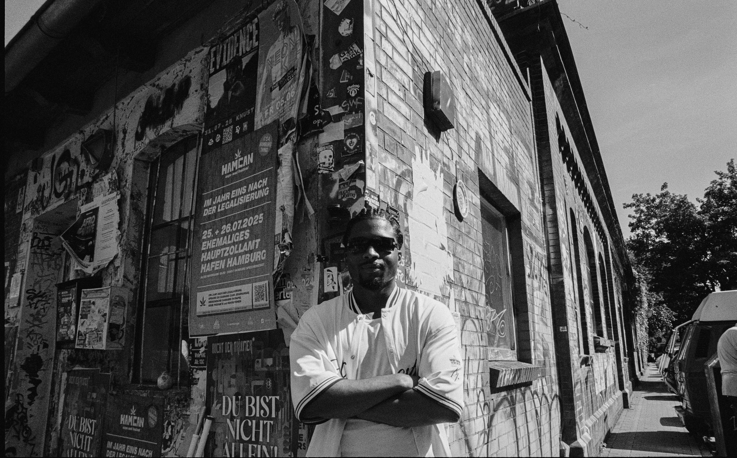 Hombre con gafas y camiseta blanca cruzando los brazos en la calle, en una zona urbana, con paredes cubiertas de carteles y grafiti, y árboles al fondo.