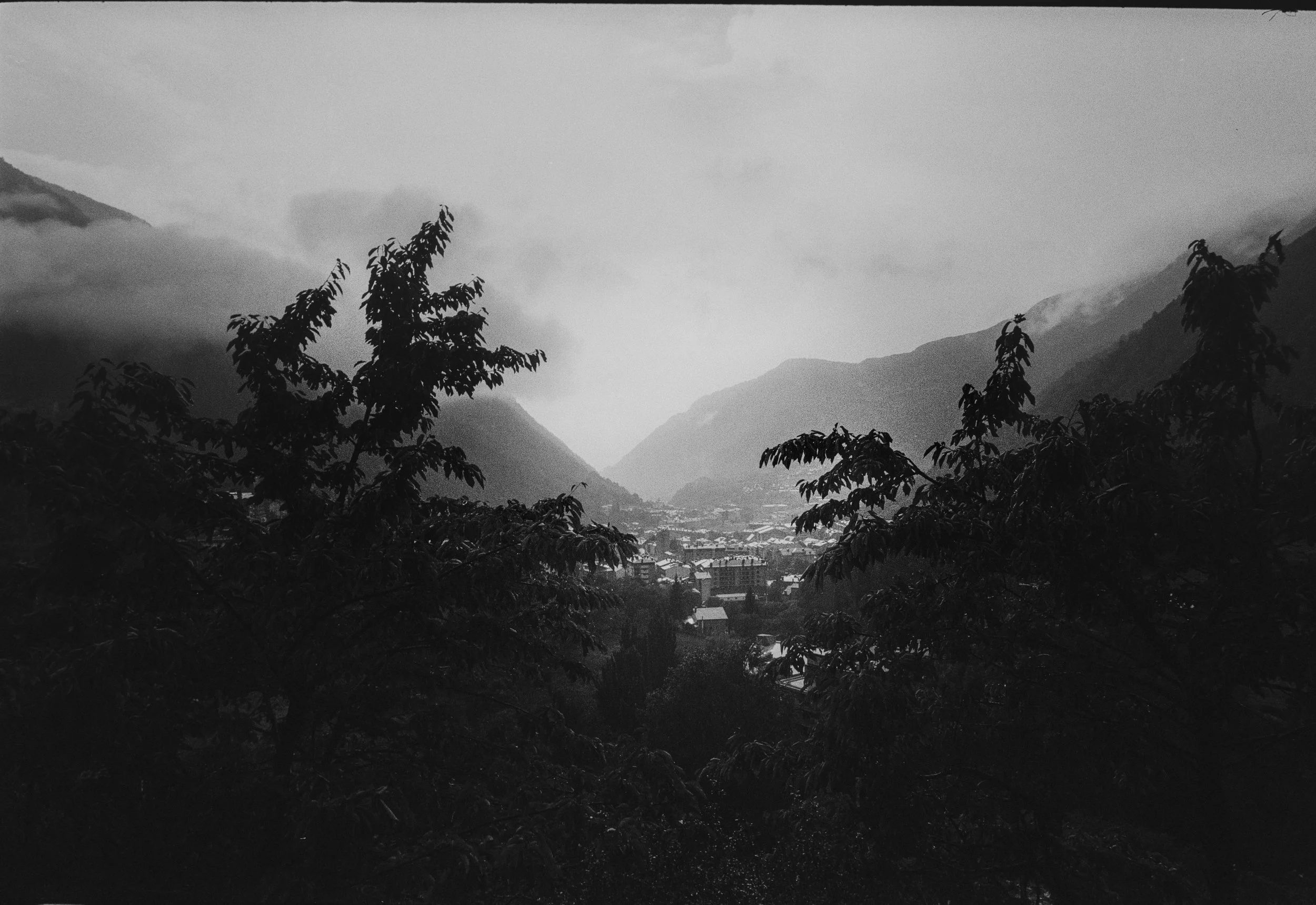 Paisaje montañoso con árboles en primer plano y ciudad en el valle, en un día nublado.