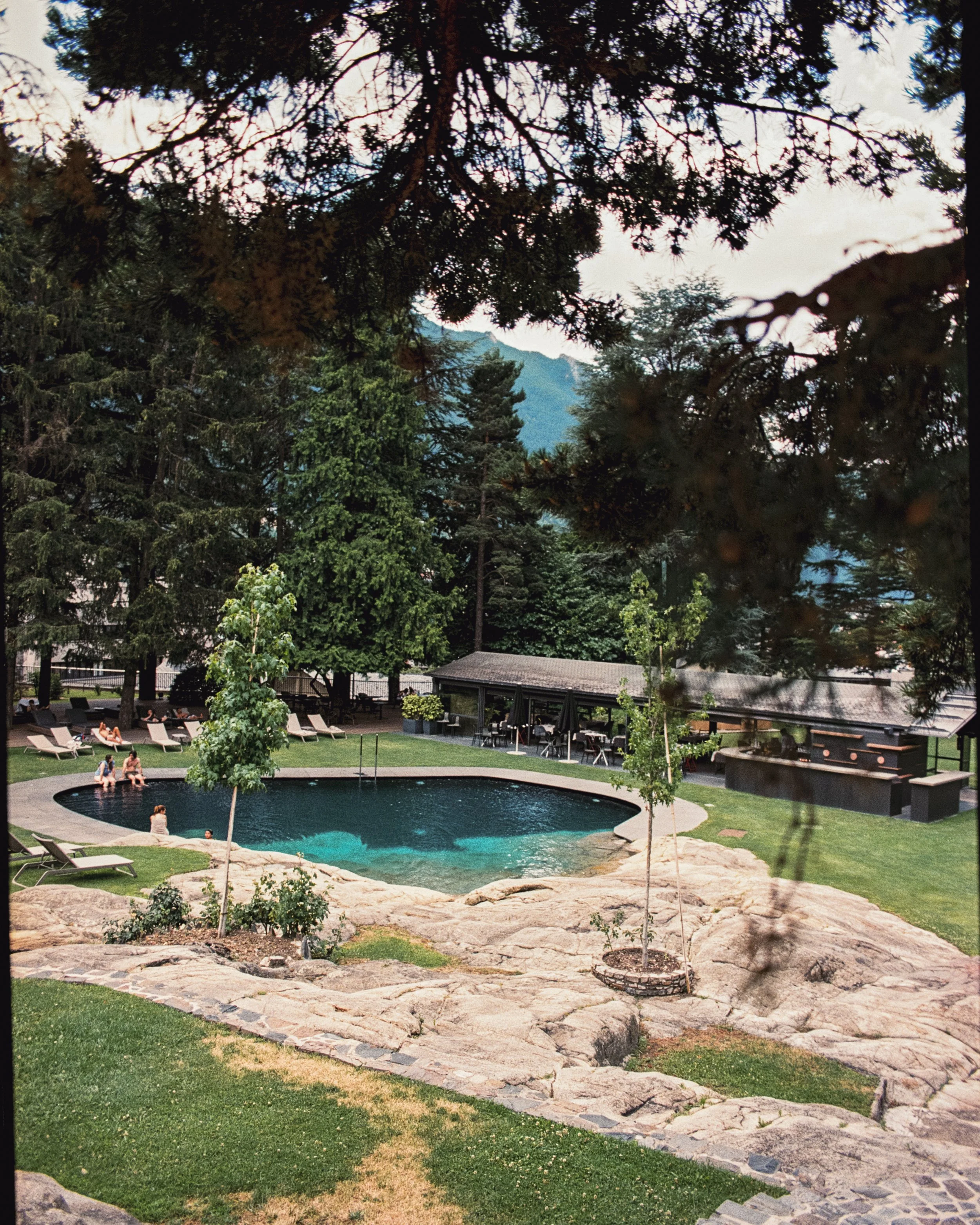 Piscina circular rodeada de piedra y sillas tumbona en un jardín con árboles y fondo de montañas.