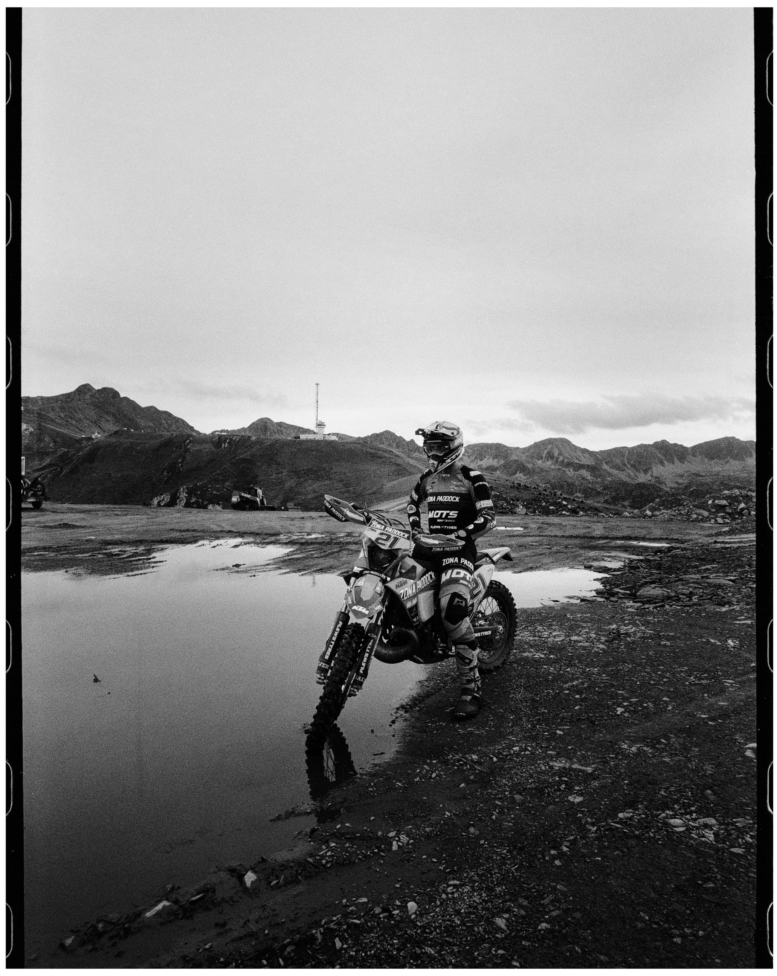 Persona con equipo de motocross en un terreno montañoso y un charco de agua, con montañas en el fondo y el cielo despejado.