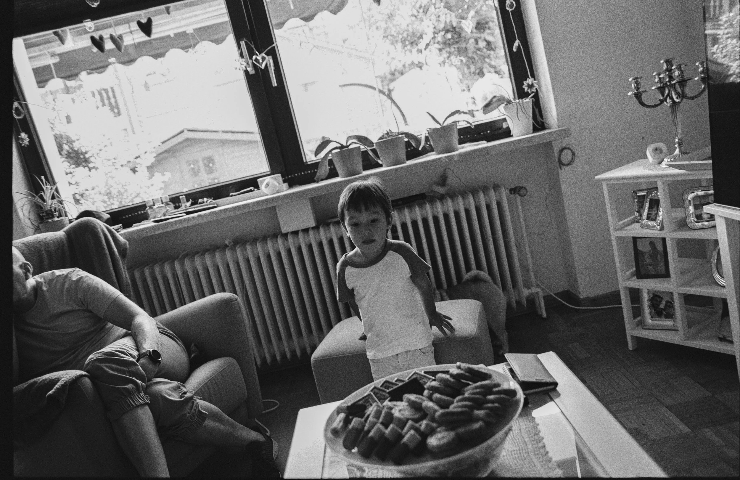 Escena en una sala de estar con niños. Una niña está sentada en un sillón y un niño está de pie frente a una mesa con dulces. La ventana grande tiene decoraciones colgantes y plantas en la ventana. Hay muebles y objetos decorativos en la habitación.