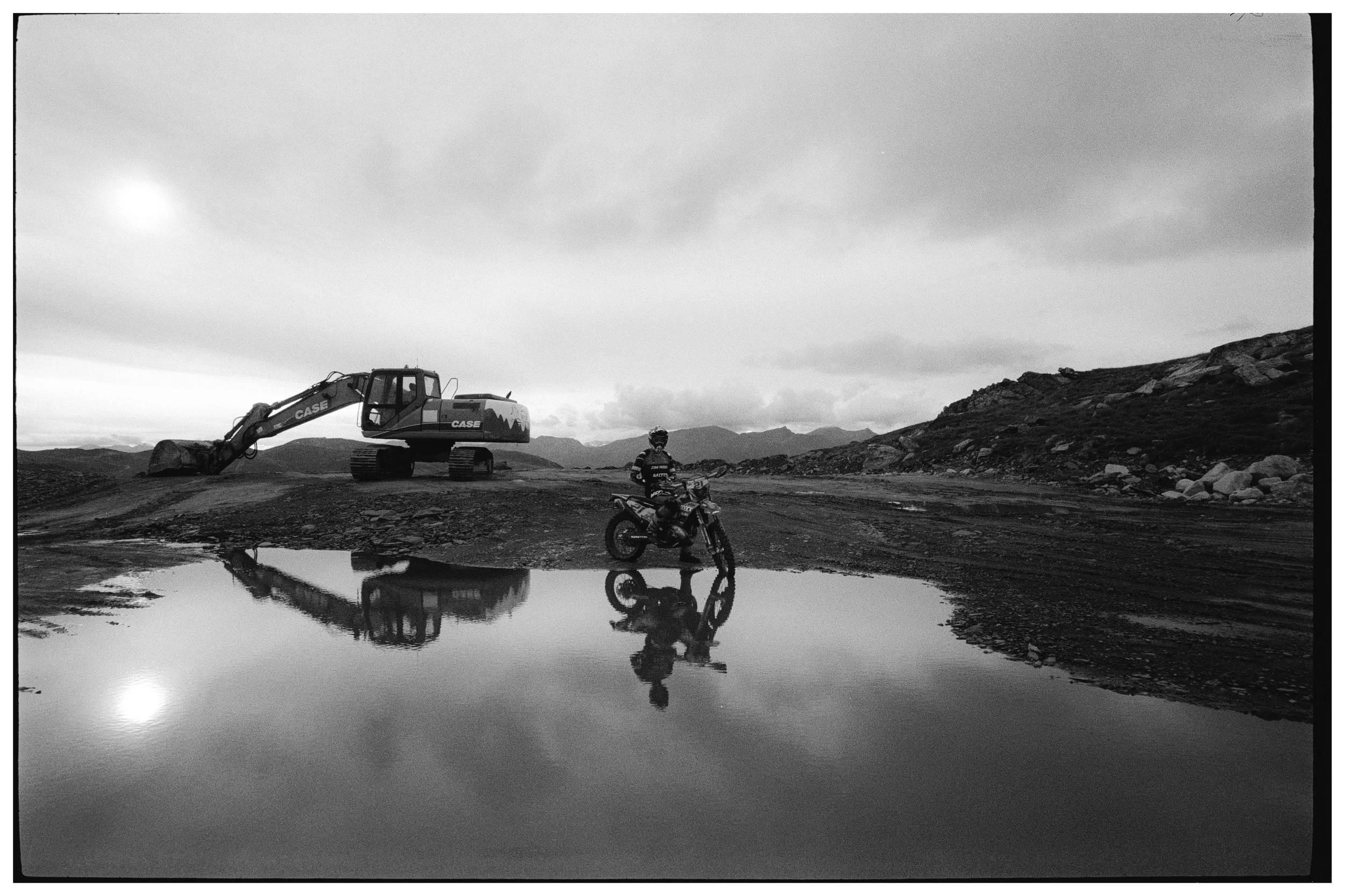 Paisaje árido con una retroexcavadora y una motocicleta en primer plano, reflejadas en un charco de agua, montañas al fondo y cielo nublado.