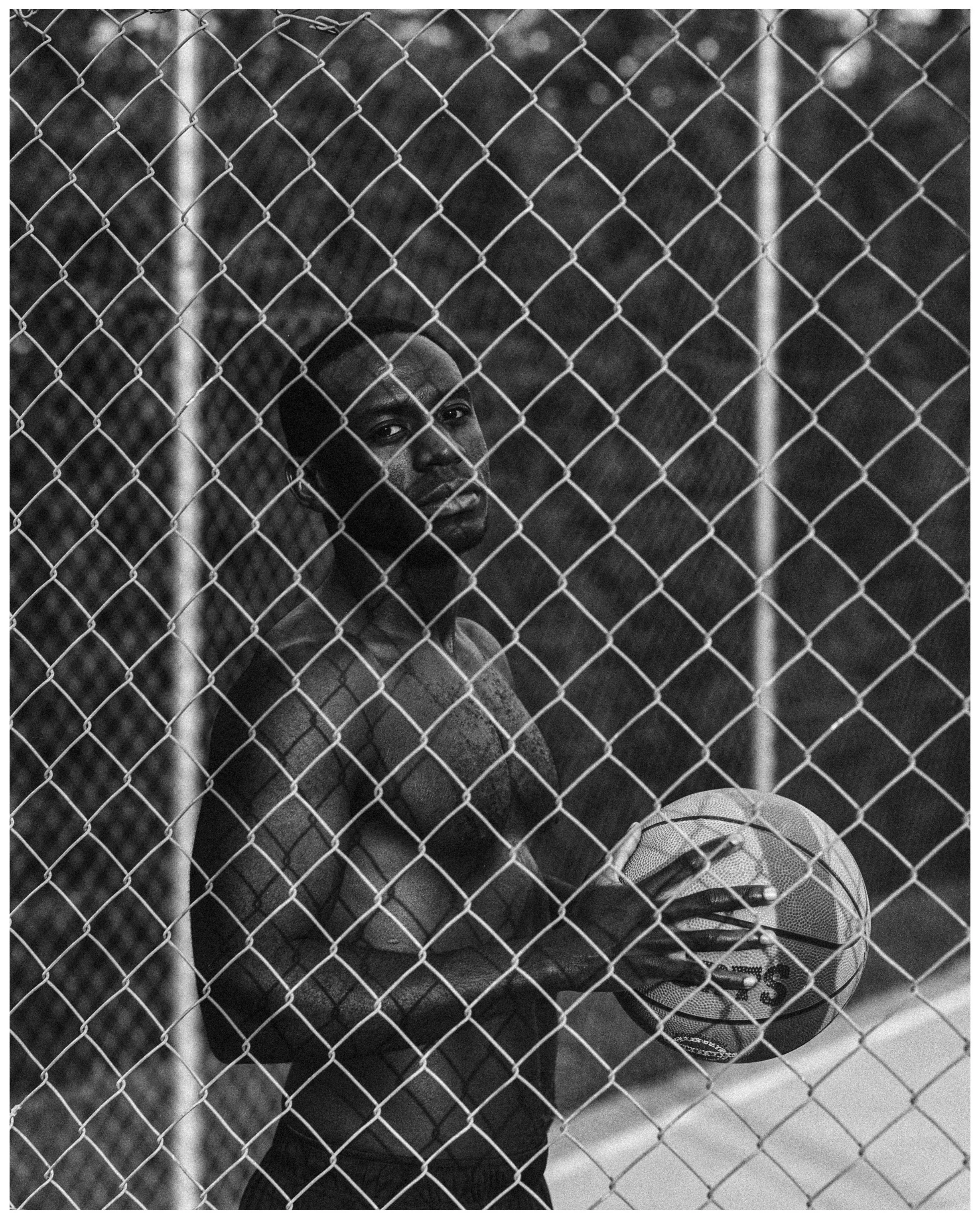 Joven afroamericano sosteniendo una pelota de baloncesto detrás de una cerca de malla metálica en un entorno al aire libre, en blanco y negro.