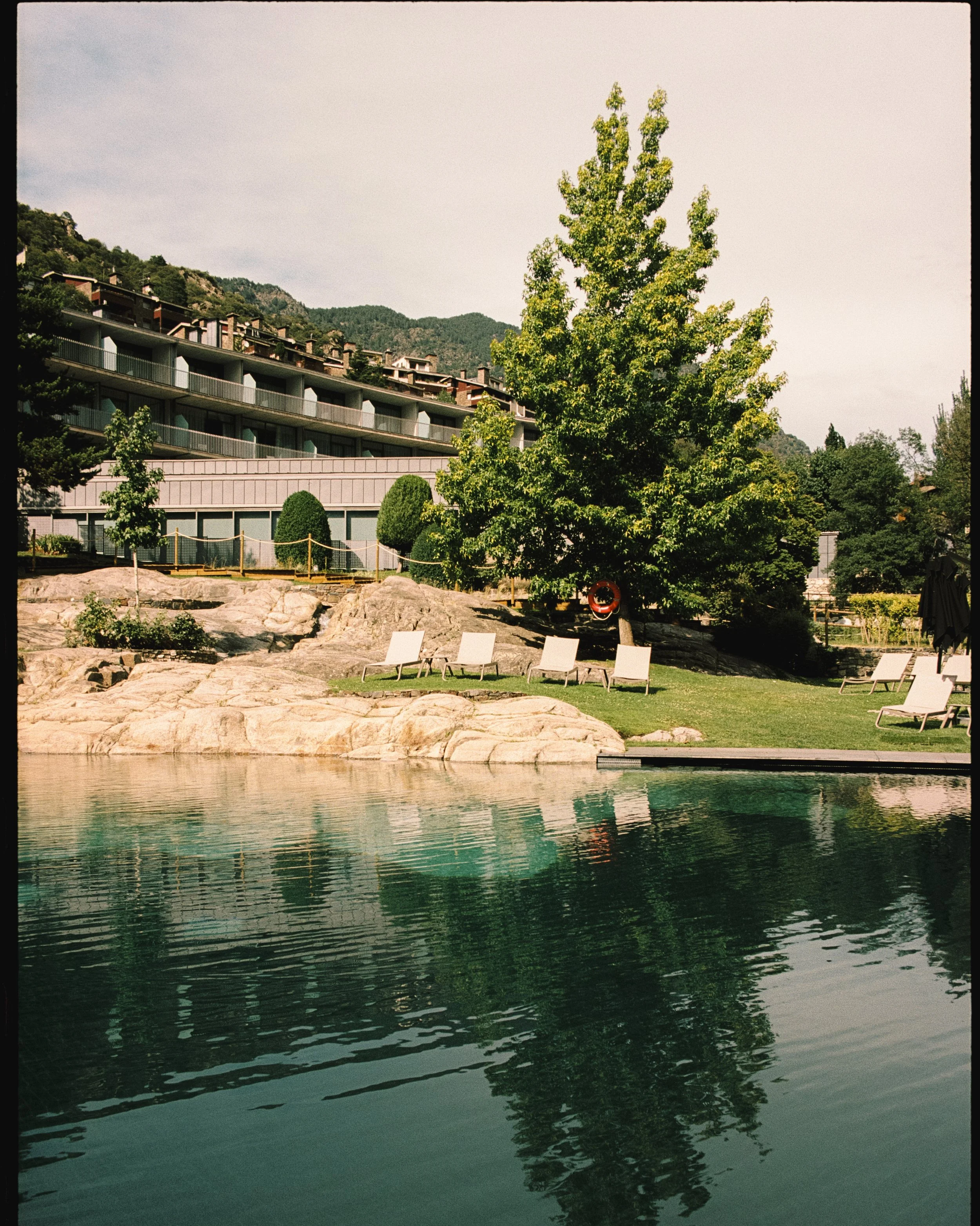 Piscina con sillas reclinables, árboles y un edificio en el fondo rodeado de montañas.