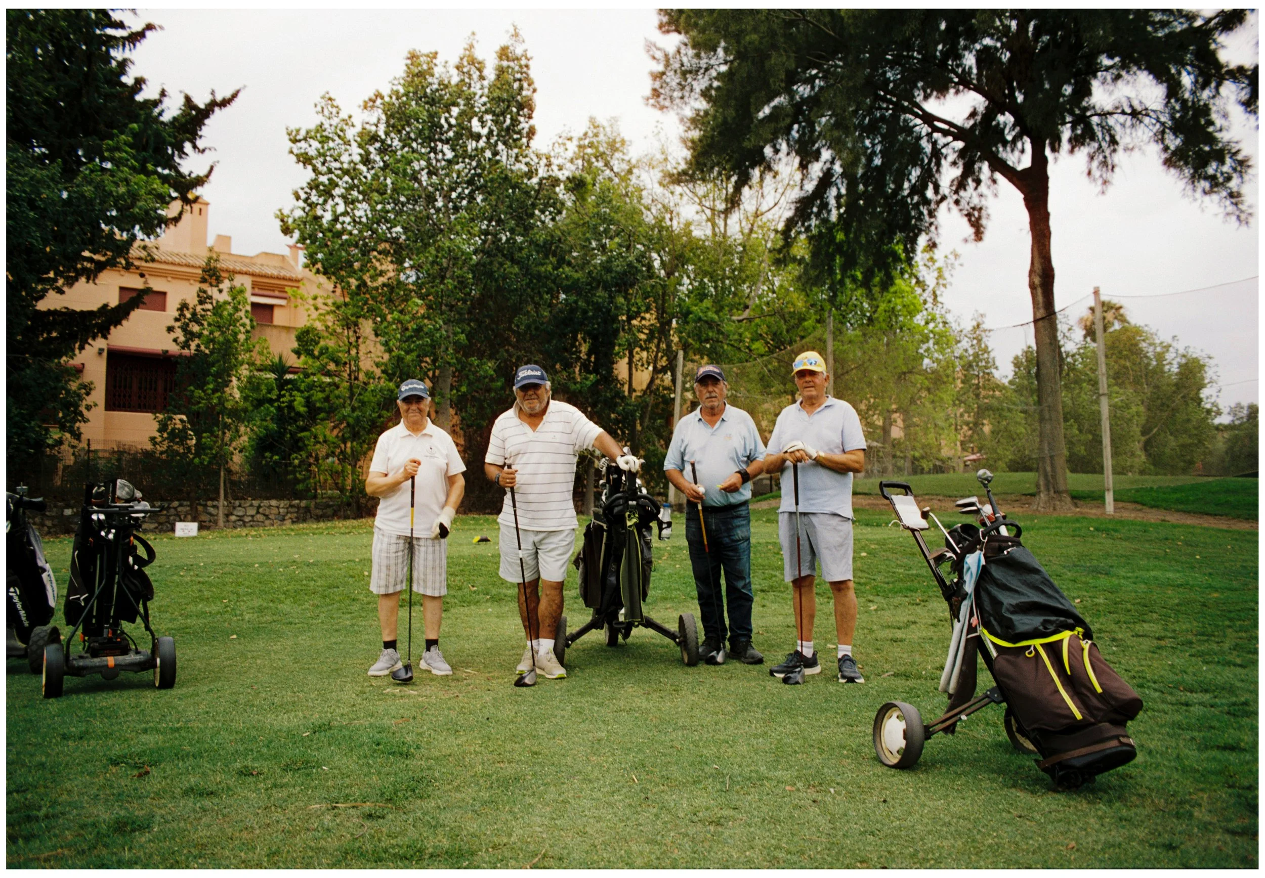 Cinco personas mayores con equipo de golf en un campo en un día soleado, rodeados de árboles y casas en el fondo.