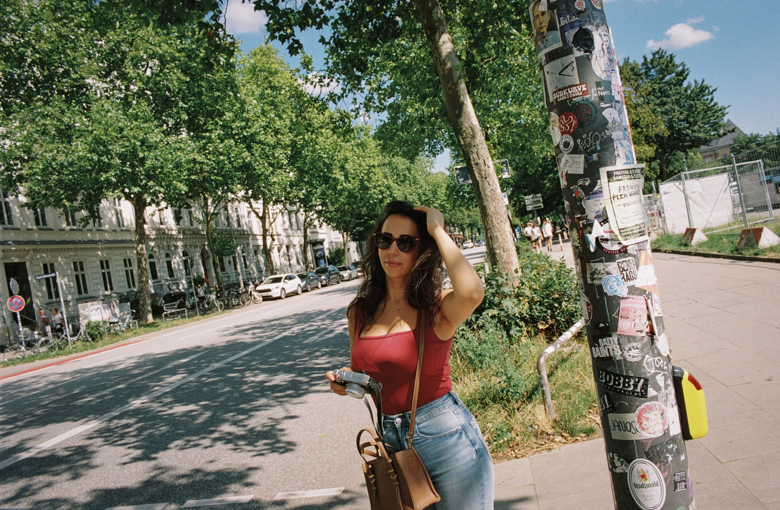 Mujer con gafas de sol posando en la acera, con árboles y edificios clásicos al fondo, sosteniendo una cámara en una mano y con bolso colgado del hombro, en un día soleado en la ciudad.