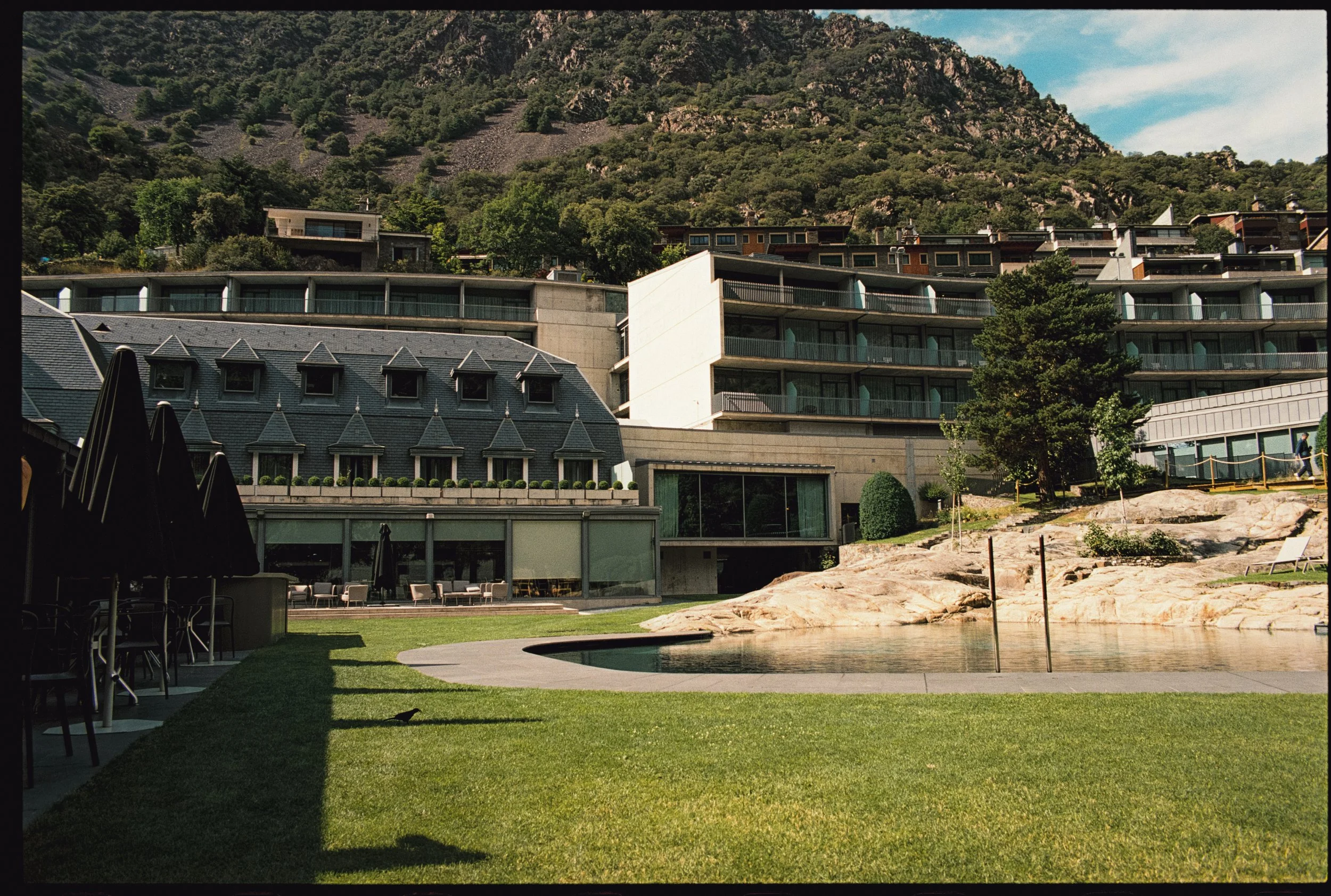 Edificio moderno con jardines y piscina, montañas y cielo azul de fondo.