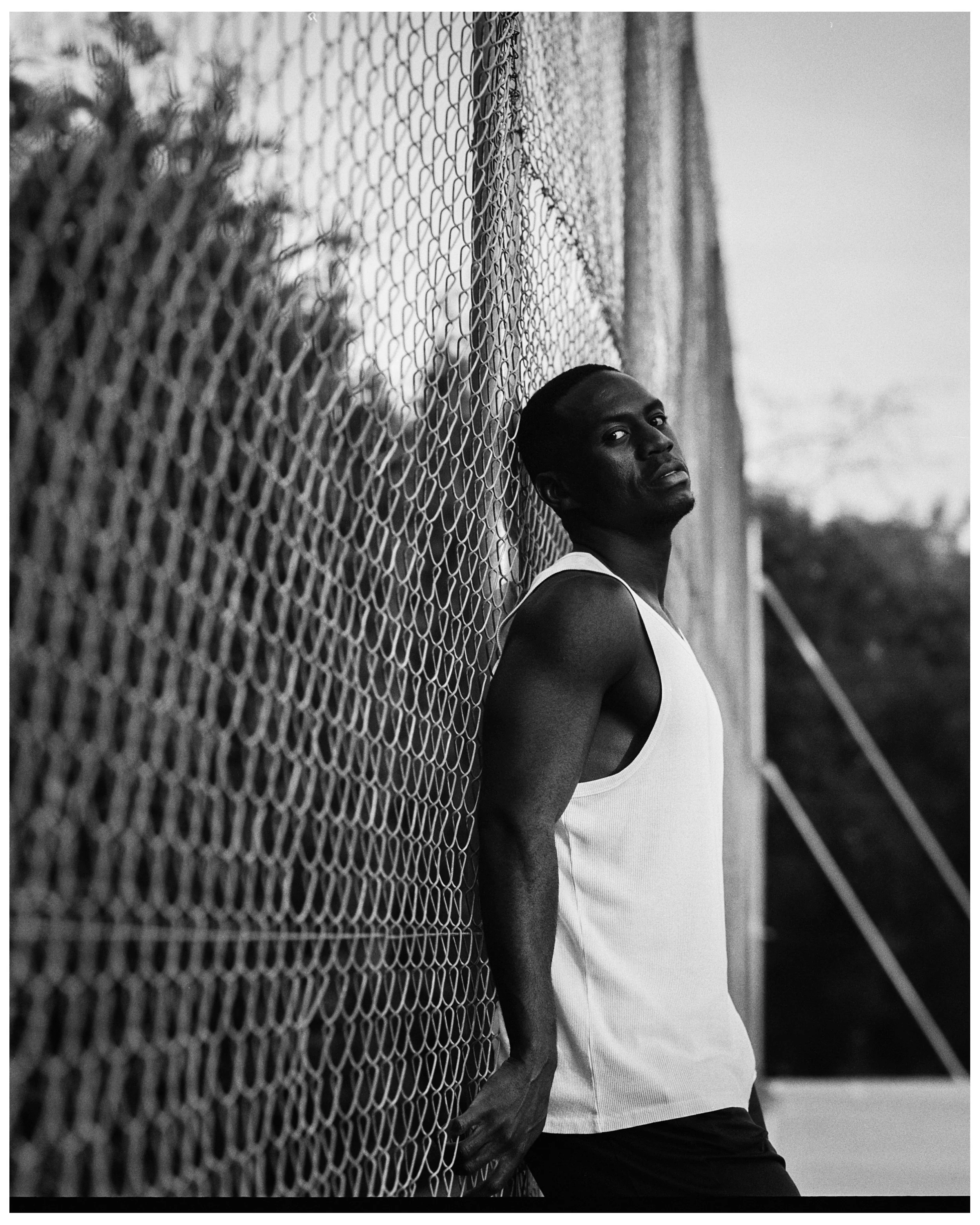 Joven hombre afroamericano de complexión atlética, con ropa deportiva blanca, apoyado de espaldas en una valla de metal en una cancha, mirando hacia la cámara en un entorno exterior en blanco y negro.