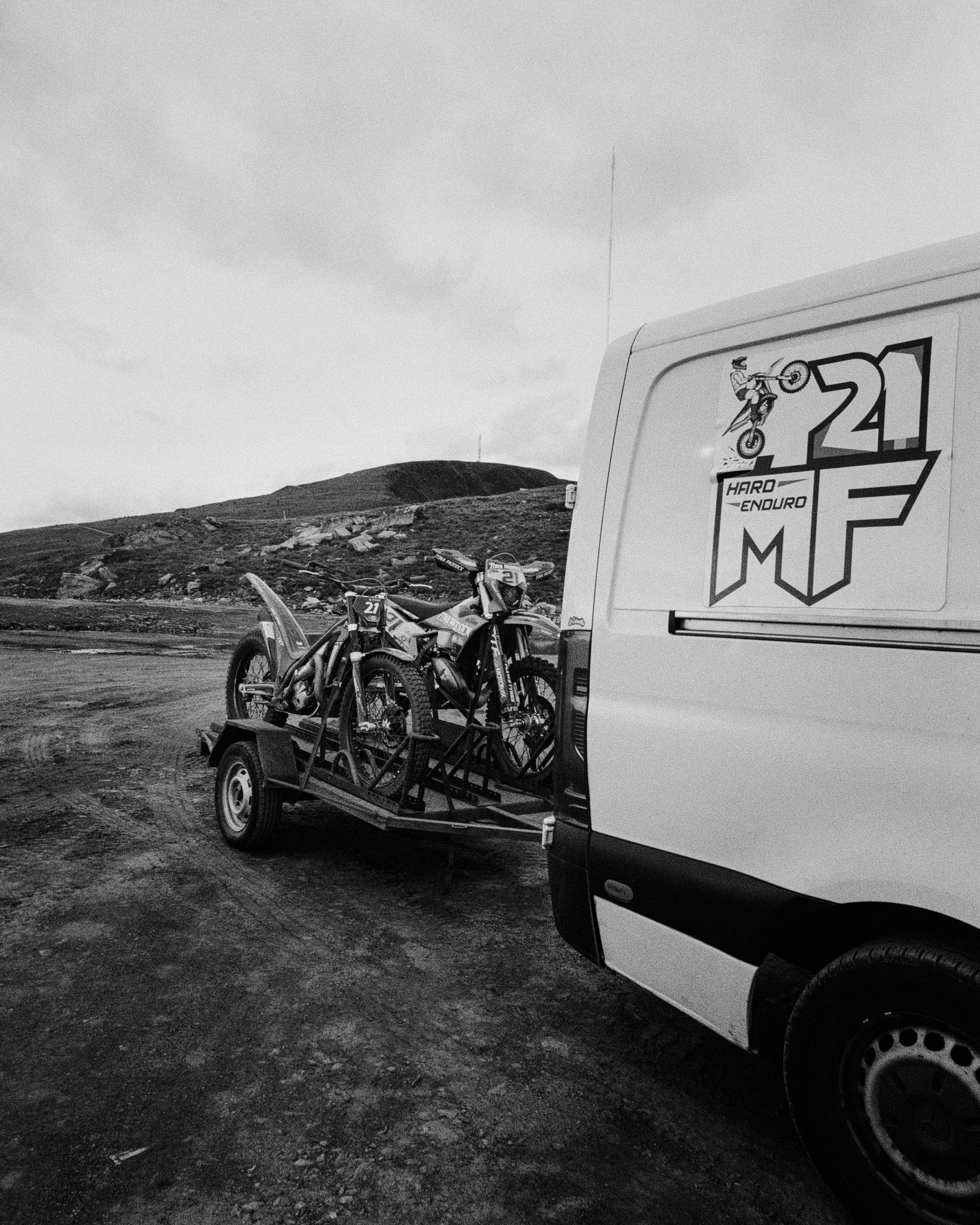 Camioneta blanca con logotipo de competición de motocross, llevando dos motos de dirt bike en remolque en un terreno montañoso.