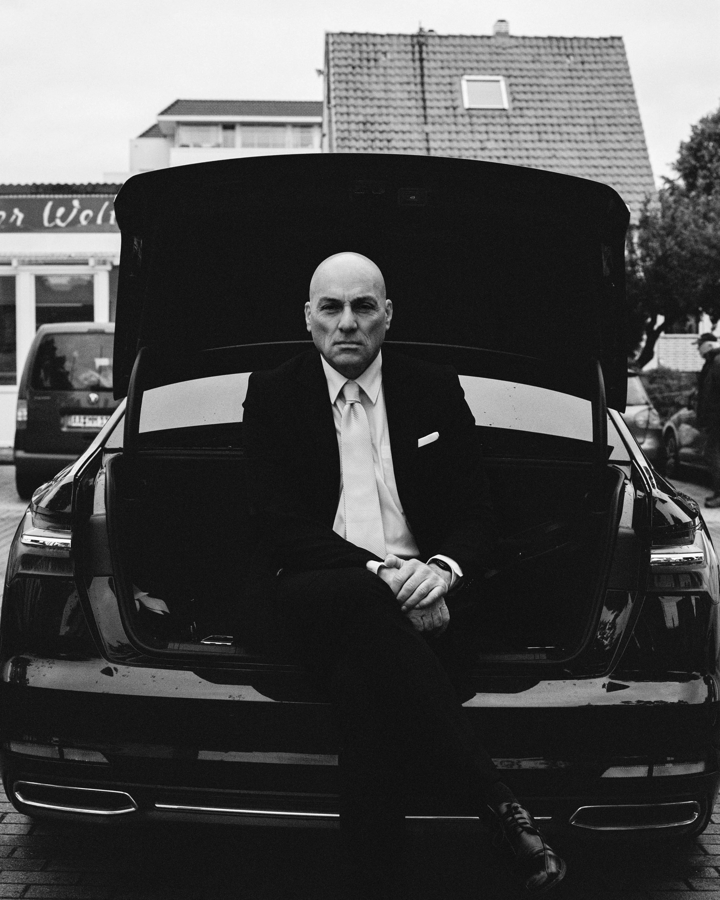 Hombre con traje y corbata sentado en la cajuela de un coche negro con capota abierta, en un entorno urbano, en blanco y negro.