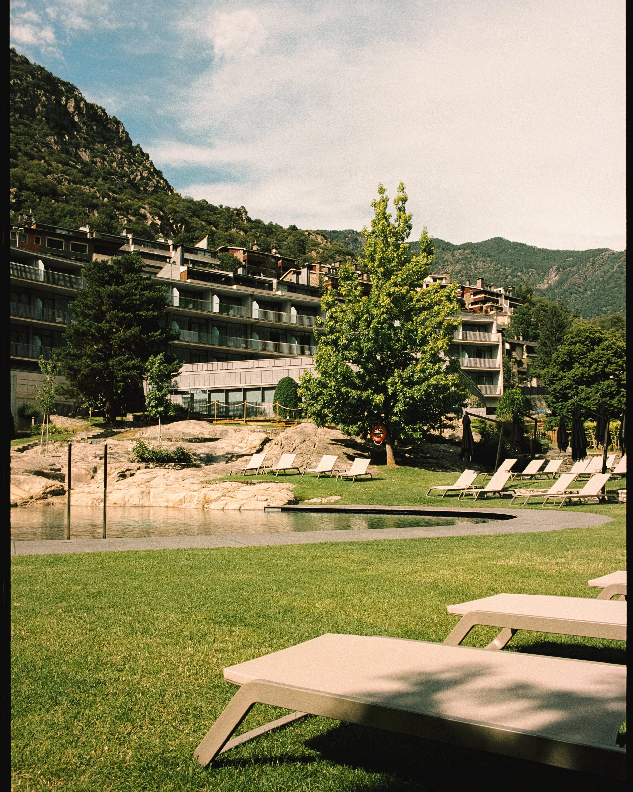 Zona de descanso con sillas de playa junto a una alberca, rodeada de césped y árboles, con edificios residenciales en las montañas al fondo y un cielo con pocas nubes.
