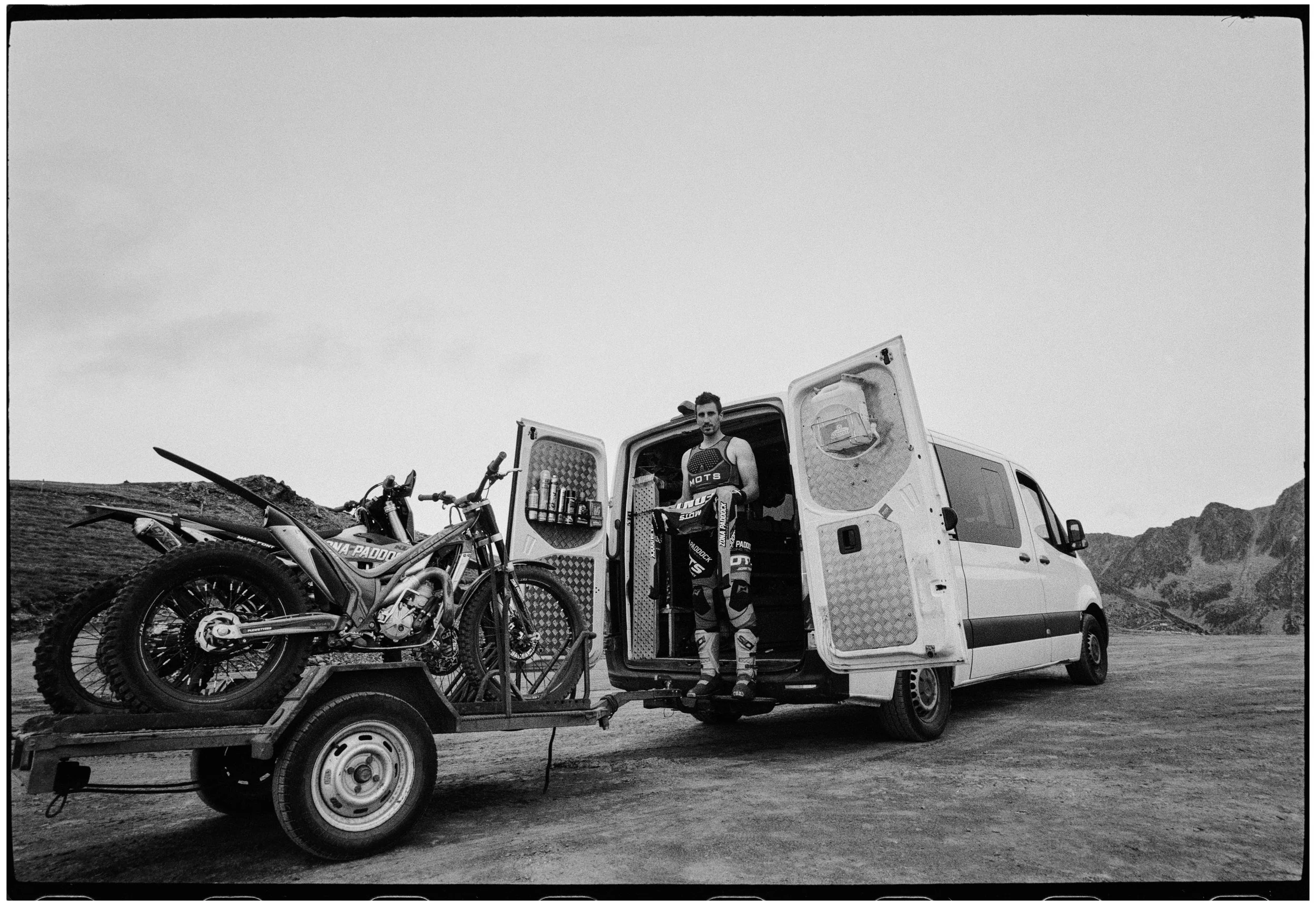 Un hombre en ropa de motocross con un casco en la mano, de pie en la parte trasera de una furgoneta blanca, cargada con una moto de dirt bike en una carreta, en un paisaje montañoso.