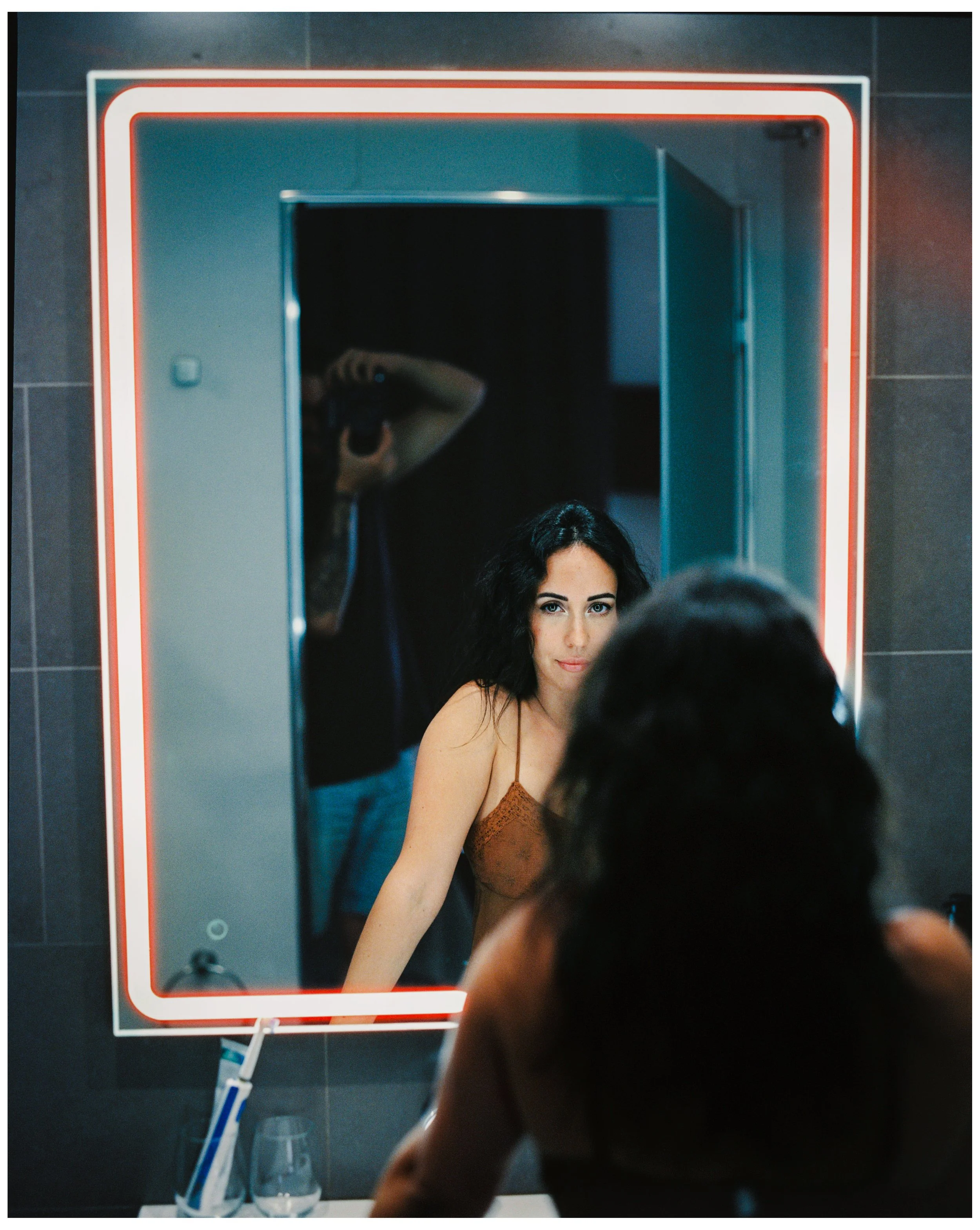 Una mujer con cabello negro y piel clara se mira en un espejo de baño con marco de luces de neón, mientras se arregla frente al espejo.