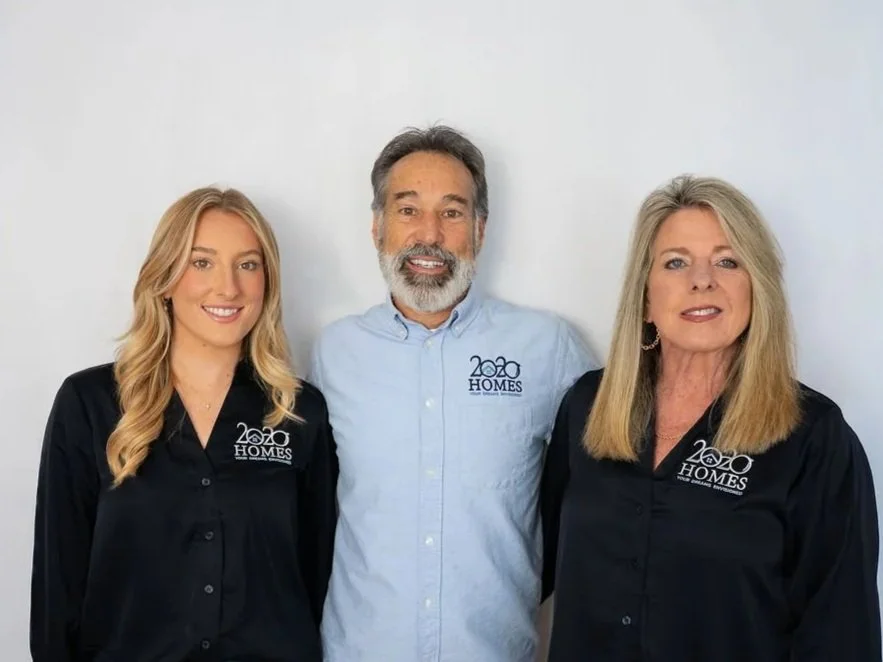 Three people standing side by side, smiling, with a plain gray background. The two women on each side are wearing black shirts with a logo that says '2020 HOMES', and the man in the middle is wearing a light blue shirt with the same logo.