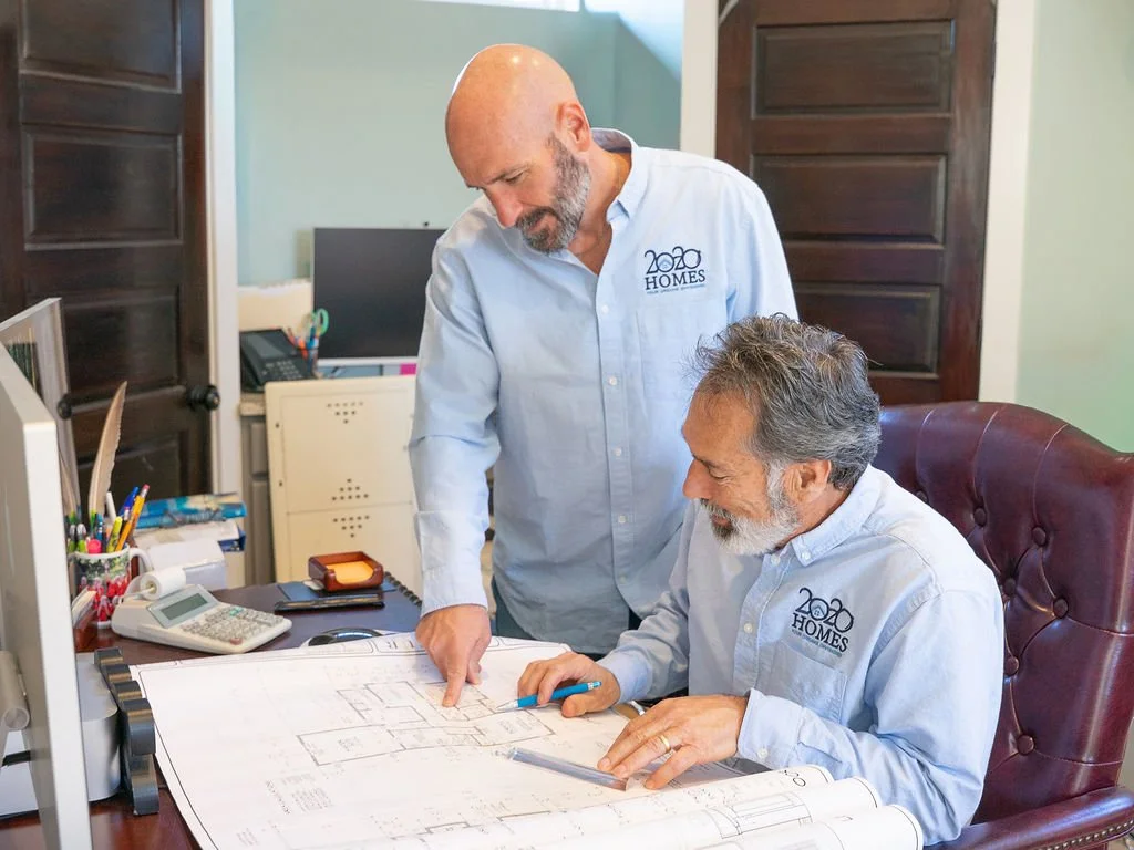 Two men wearing light blue shirts with a 2020 HOMES logo, working on architectural plans at a desk in an office. One man is sitting at the desk, holding a pencil, while the other is standing and pointing at the plans.