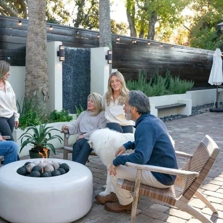 Group of five people sitting around a fire pit in an outdoor patio, with one person petting a large white dog. The setting features a  water fountain, trees, and string lights.