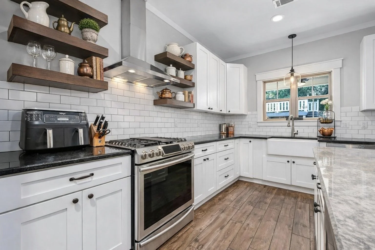 Kitchen with white cabinets, black countertops, stainless steel stove, white subway tile backsplash, open wooden shelves with dishes and decor, window over sink, and wooden floors.