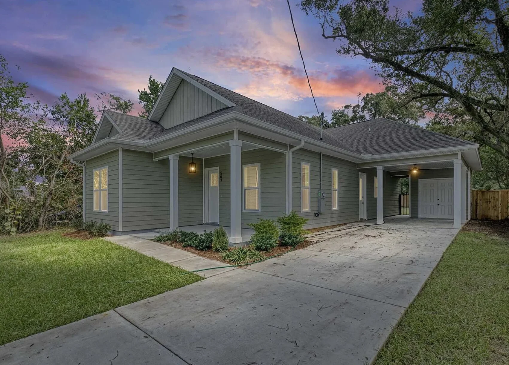 A modern gray house with a covered front porch and a garage, set against a colorful sunset sky with trees in the background.
