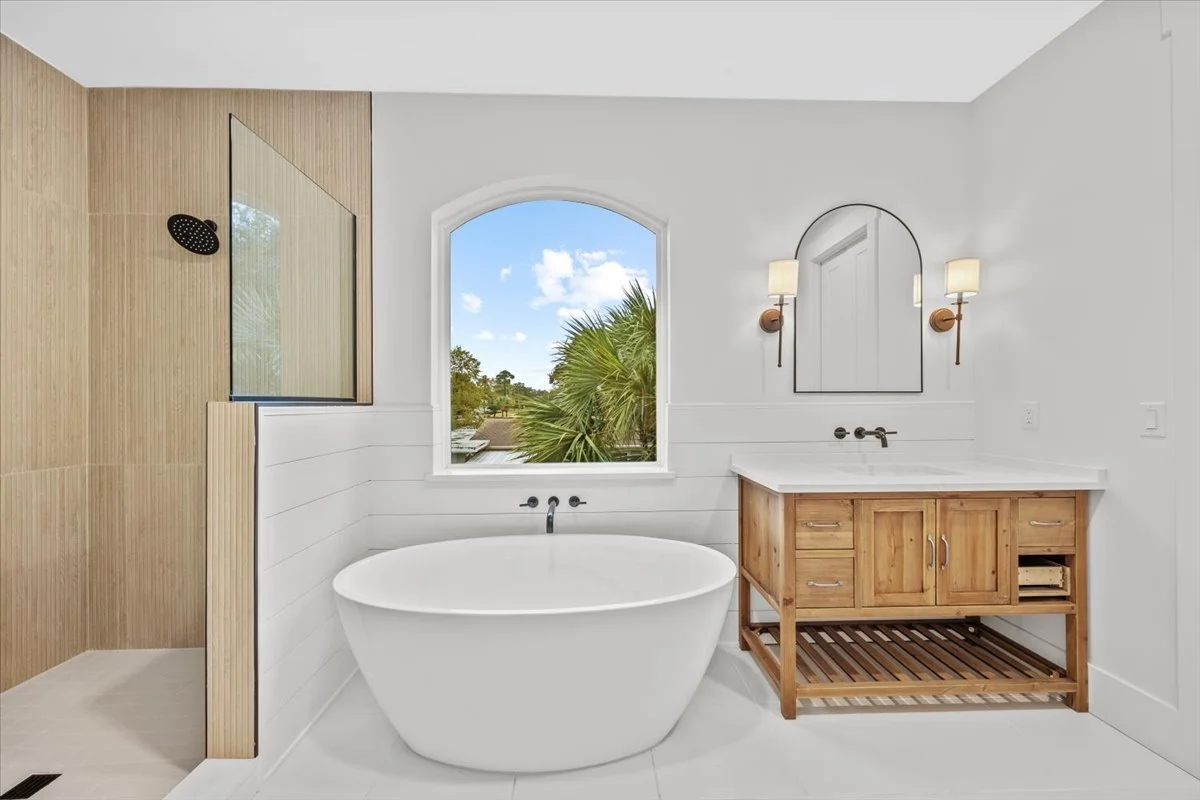 Modern bathroom with a freestanding bathtub beneath a window, wooden vanity with a white countertop, oval mirror, and wall-mounted lights. A walk-in shower with wooden paneling is on the left.