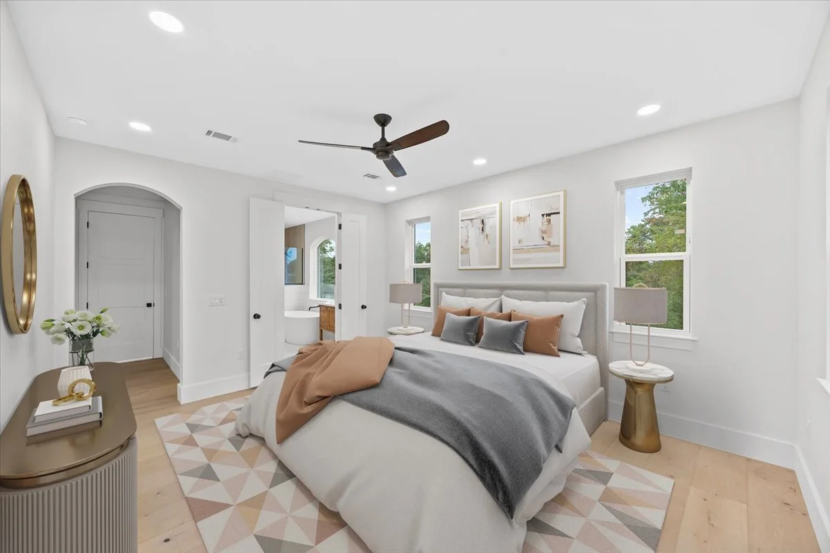 Bright, modern bedroom with white walls, large bed with multiple pillows, two side tables with lamps, a ceiling fan, and three abstract artwork pieces above the bed. Natural light from two windows, hardwood floors, and a geometric area rug.