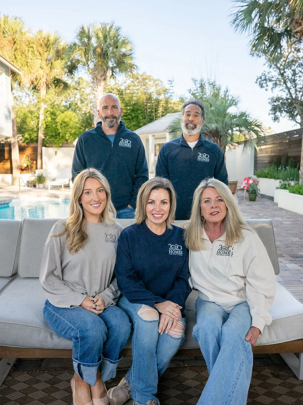Group of five people, three women and two men, sitting and standing outdoors near a pool in a backyard with palm trees and potted plants, all wearing jackets with a logo.