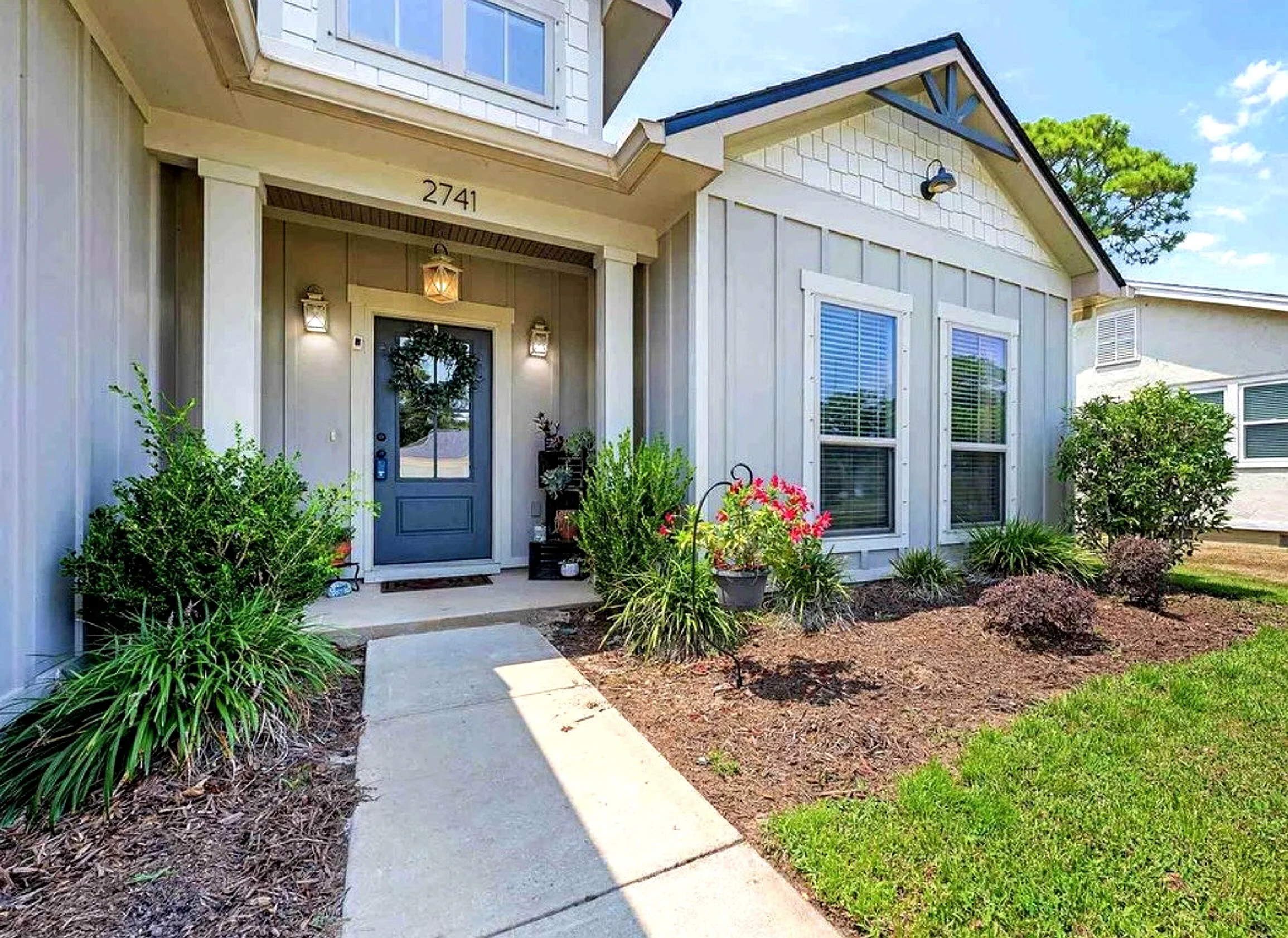 Front yard of a house with a concrete walkway, green shrubs, flowering plants, and a blue front door under a porch with a lantern and lights. The house has light-colored siding and two windows with blinds.