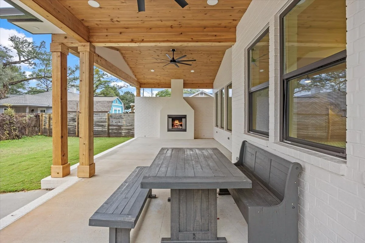 Covered outdoor patio with a concrete floor, a gray wooden dining set and bench, white brick wall with multiple windows, and a built-in fireplace with a fire burning. The roof is wood-paneled with a black ceiling fan, and the area is surrounded by a grassy yard with a wooden fence and trees in the background.