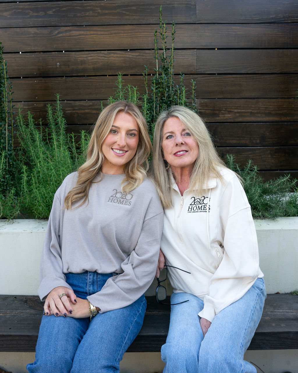 Two women sitting on a bench in front of a wooden wall and green plants, both wearing casual clothing with '2020 Homes' logos.