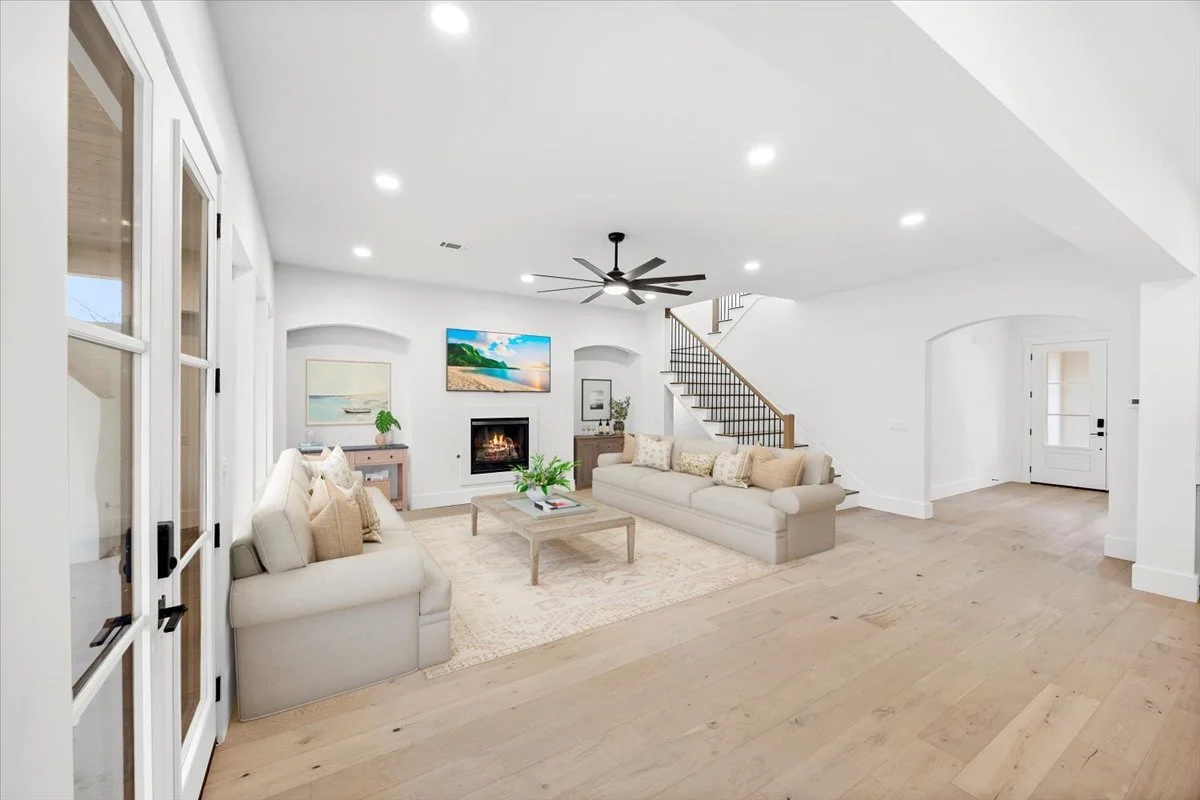 Bright living room with white walls and ceiling, wooden floor, two beige sofas with pillows, a coffee table, a fireplace with a TV above it, a staircase with wooden handrails, large glass doors on the left, and a door in the background.