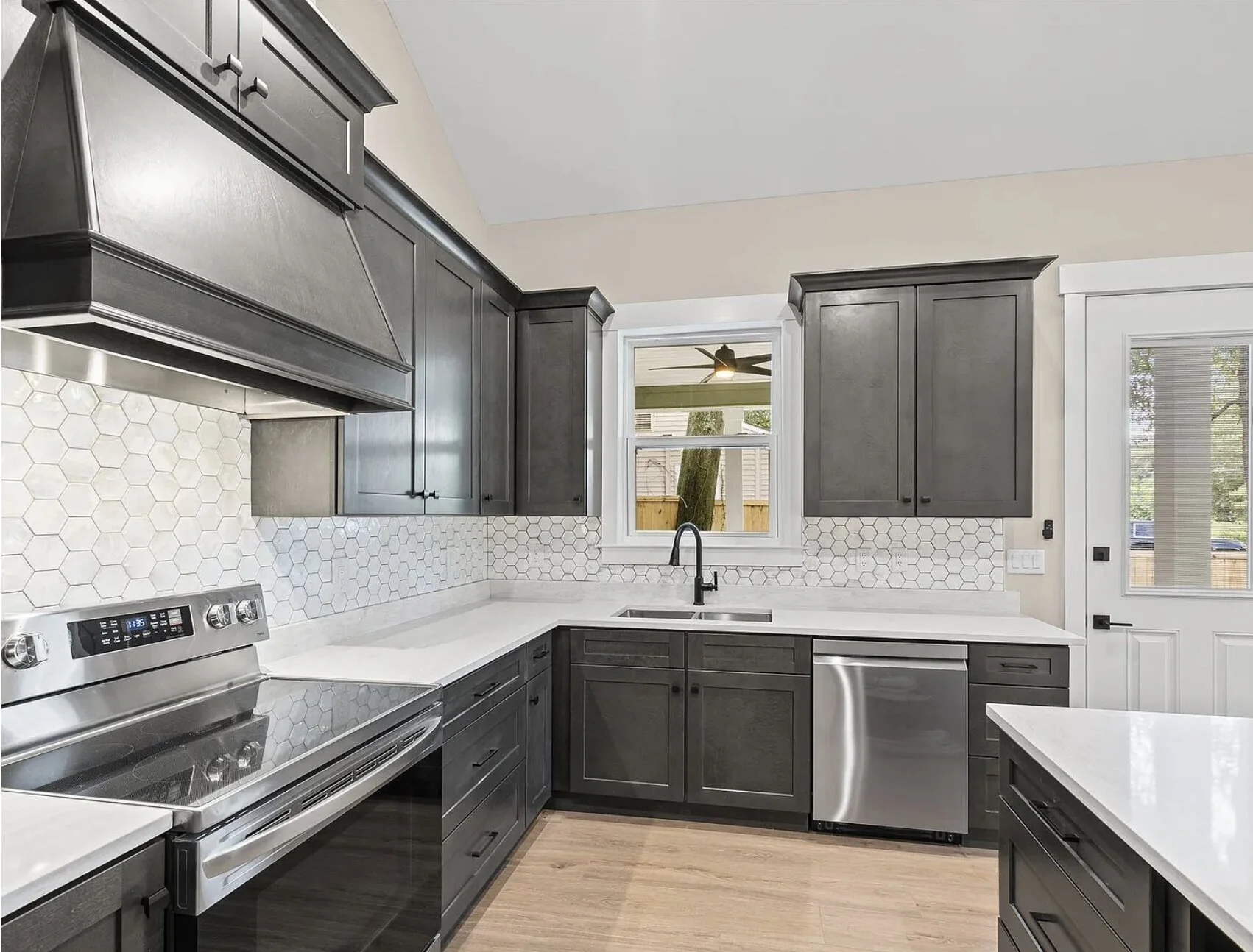 Modern kitchen with dark cabinets, white countertops, hexagonal tile backsplash, stainless steel appliances, a window, and a door leading outside.