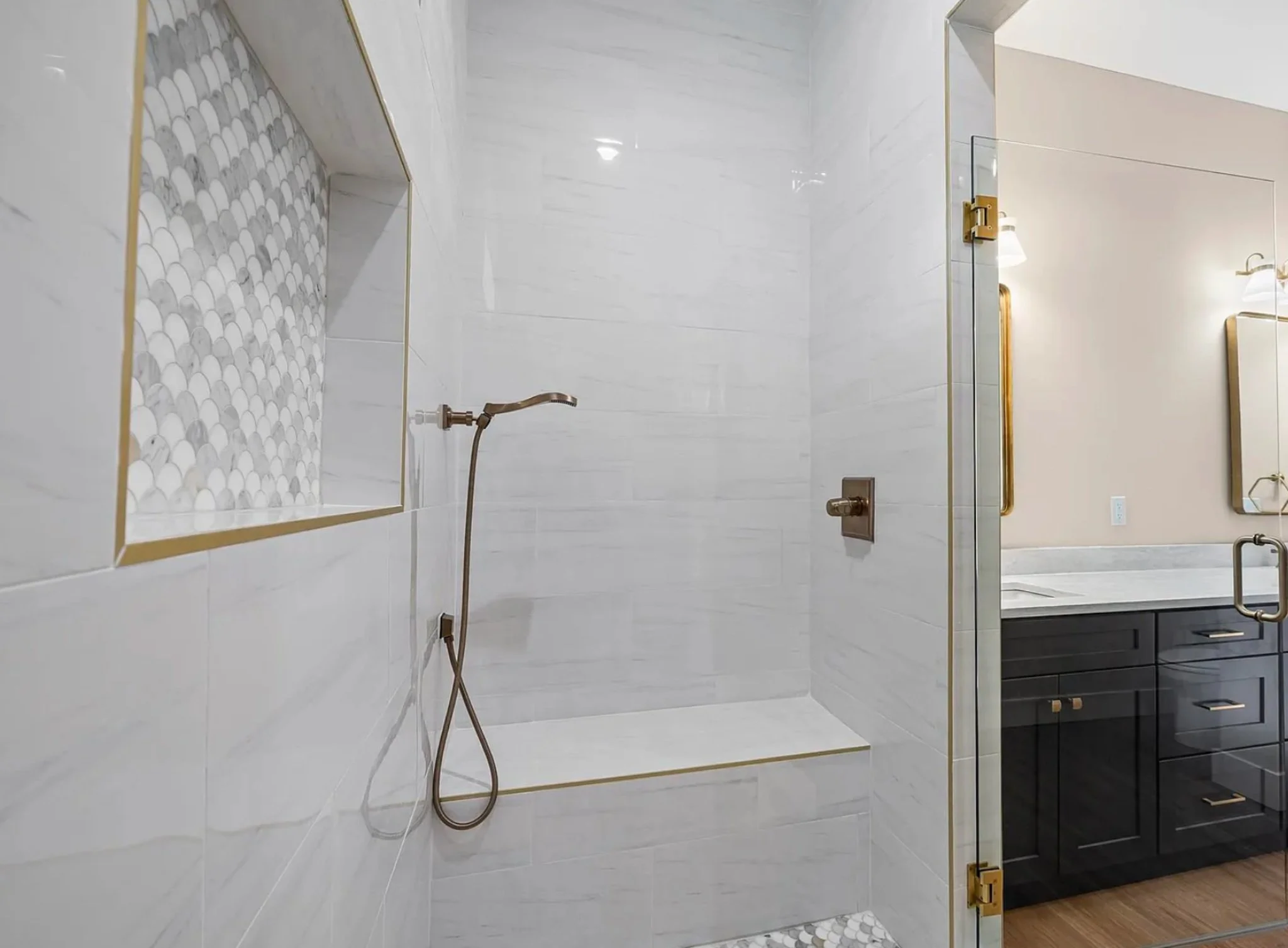A modern minimalist shower with white marble walls, a built-in niche with pebble mosaic tiles, a bronze hand-held shower head, and a marble bench. Adjacent bathroom with dark blue vanity, marble countertop, and mirrors with gold trim.