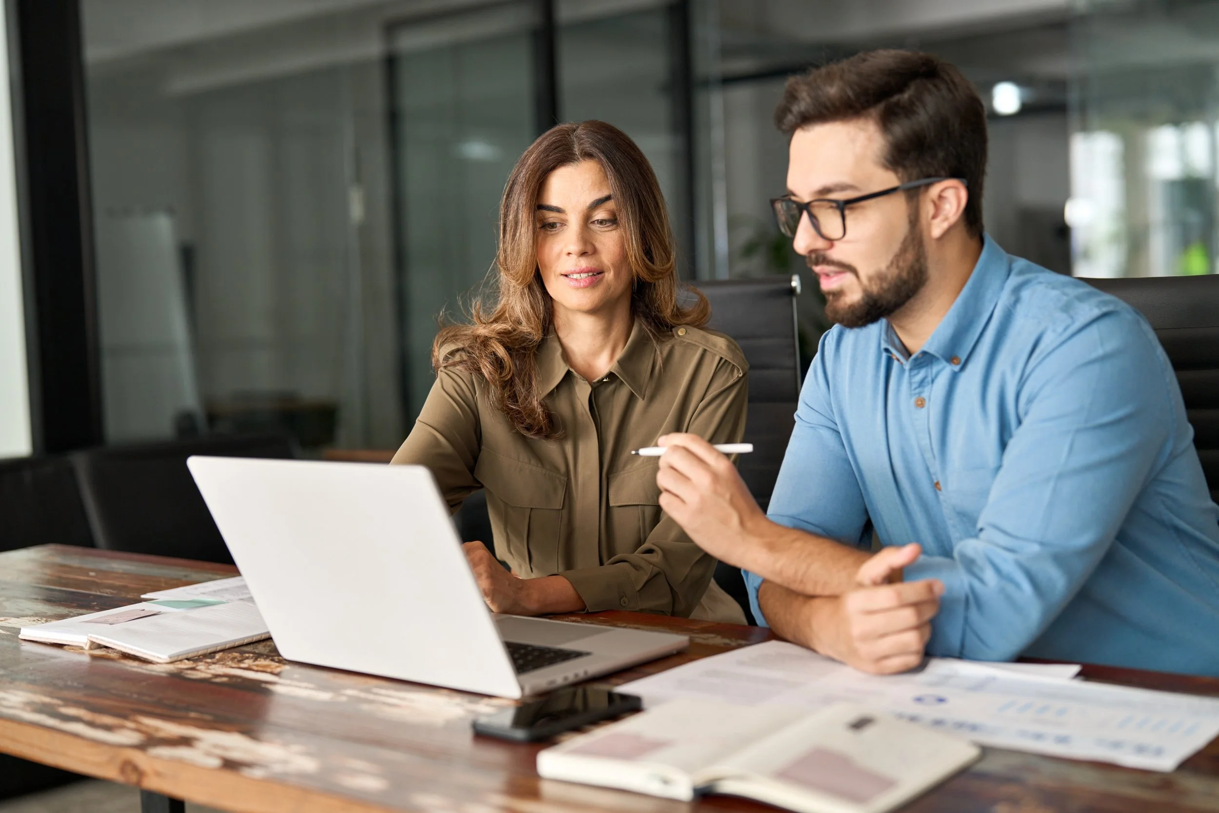 A man and a woman sitting at a desk in a modern office, looking at a laptop screen with documents and notebooks in front of them.